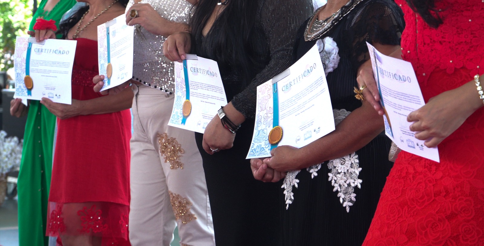 Foto colorida mostra um grupo de mulheres está posicionado lado a lado segurando certificados de conclusão do curso. Elas vestem roupas festivas em diferentes cores, como vermelho, preto e bege, e exibem os documentos com selo e fita azul.