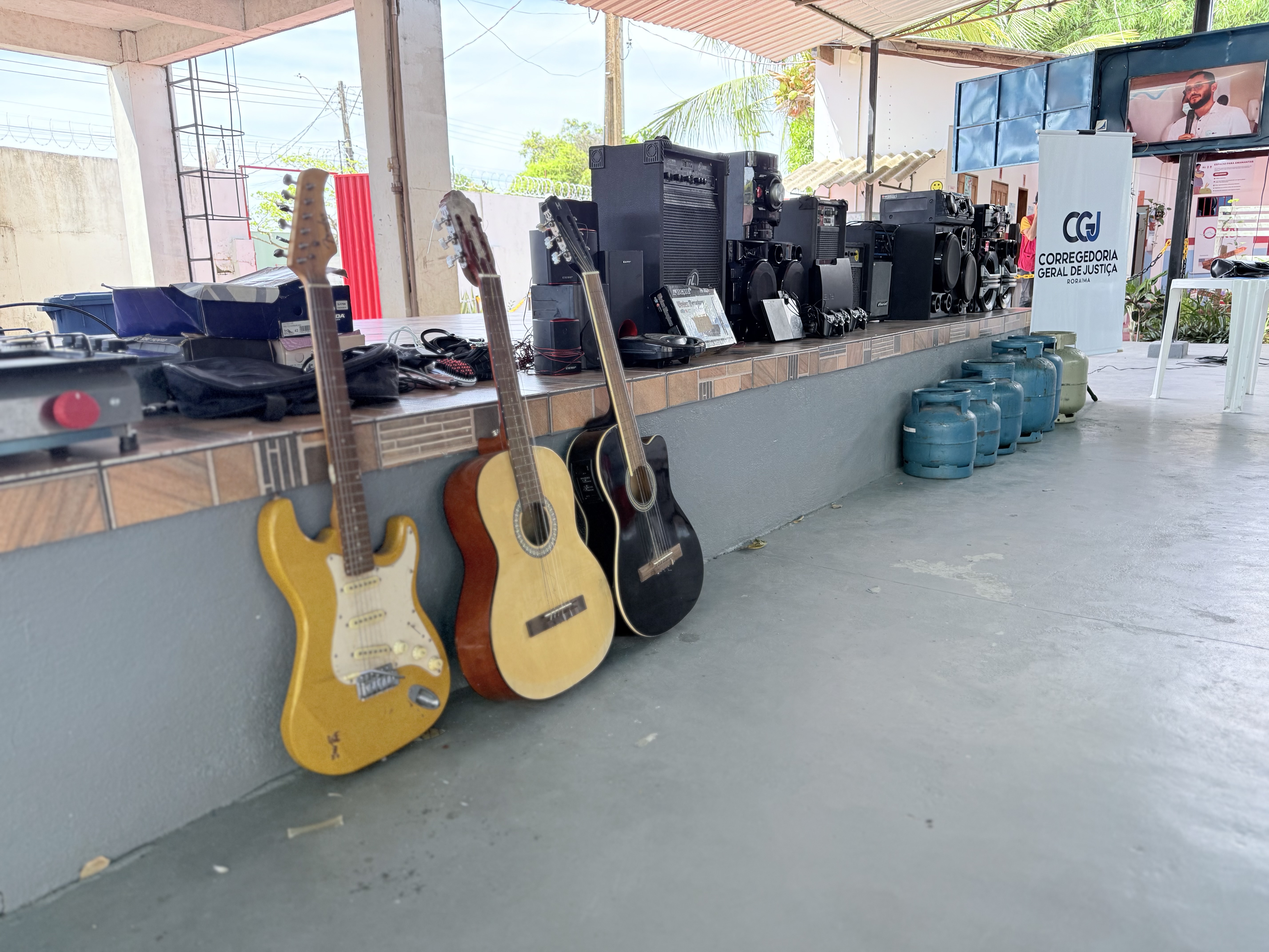 Pátio coberto com diversos equipamentos eletrônicos e instrumentos musicais sobre e ao lado de uma mureta. Uma guitarra elétrica (amarela), dois violões (amarelo e preto) e várias caixas de som pretas. Ao fundo, um banner roll-up com o logo da Corregedoria-Geral de Justiça (CGJ) e uma TV suspensa transmitem um vídeo. No chão, ao lado da mureta, estão alinhados cinco botijões de gás azul-petróleo e um cinza.