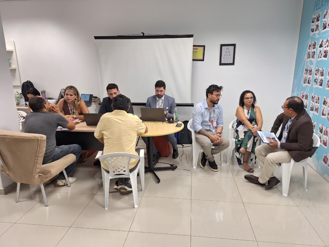 Reunião em grupo com oito pessoas sentadas ao redor de uma mesa, discutindo e trabalhando em laptops em ambiente de escritório. Ao fundo, uma tela de apresentação e uma parede decorada com fotos.