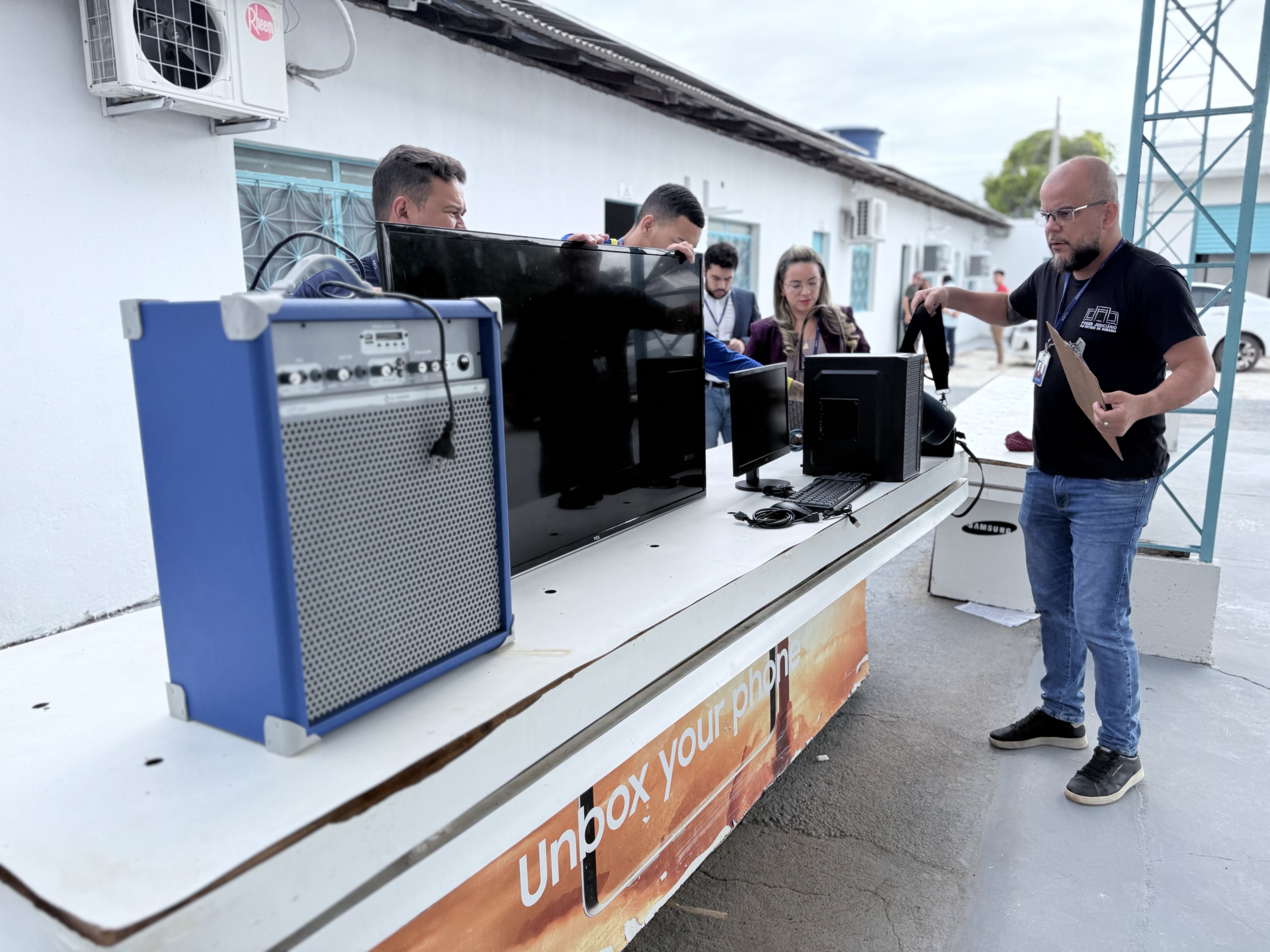  Em um pátio externo, diversos aparelhos eletrônicos (computador, televisor e caixas de som) estão organizados sobre uma mesa branca comprida. Pessoas manuseiam os itens enquanto um homem em destaque segura uma prancheta.