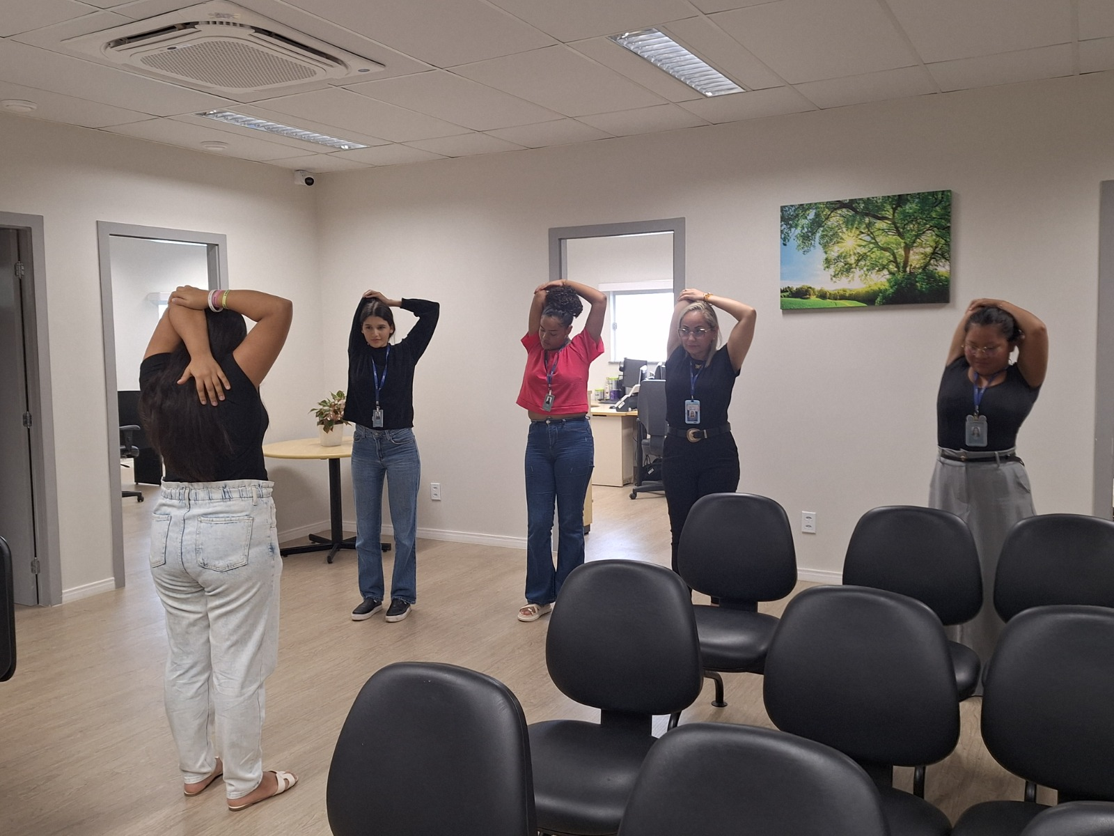 Em uma sala, cinco mulheres realizam alongamento com os braços elevados sobre a cabeça. Elas estão em pé, alinhadas lado a lado, usando crachá e roupas casuais. Ao fundo, há portas, mesa de apoio com planta e um quadro decorativo na parede. Em uma sala, cinco mulheres realizam alongamento com os braços elevados sobre a cabeça. Elas estão em pé, alinhadas lado a lado, usando crachá e roupas casuais. Ao fundo, há portas, mesa de apoio com planta e um quadro decorativo na parede.