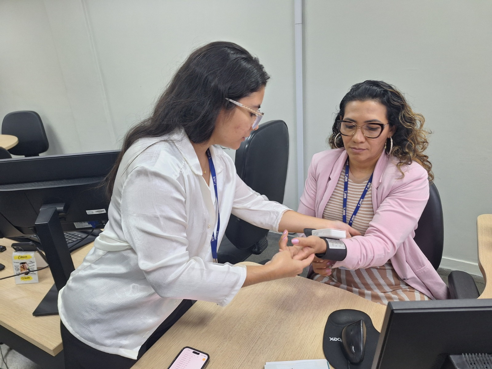 Em um ambiente de escritório, uma profissional mede a pressão arterial de uma mulher sentada. A participante usa um aparelho no braço direito enquanto a profissional segura o pulso e observa o equipamento. Ambas utilizam crachá e óculos. Sobre a mesa há computador, mouse, celular e documentos. O espaço é claro, com cadeiras e divisórias. Em um ambiente de escritório, uma profissional mede a pressão arterial de uma mulher sentada. A participante usa um aparelho no braço direito enquanto a profissional segura o pulso e observa o equipamento. Ambas utilizam crachá e óculos. Sobre a mesa há computador, mouse, celular e documentos. O espaço é claro, com cadeiras e divisórias.