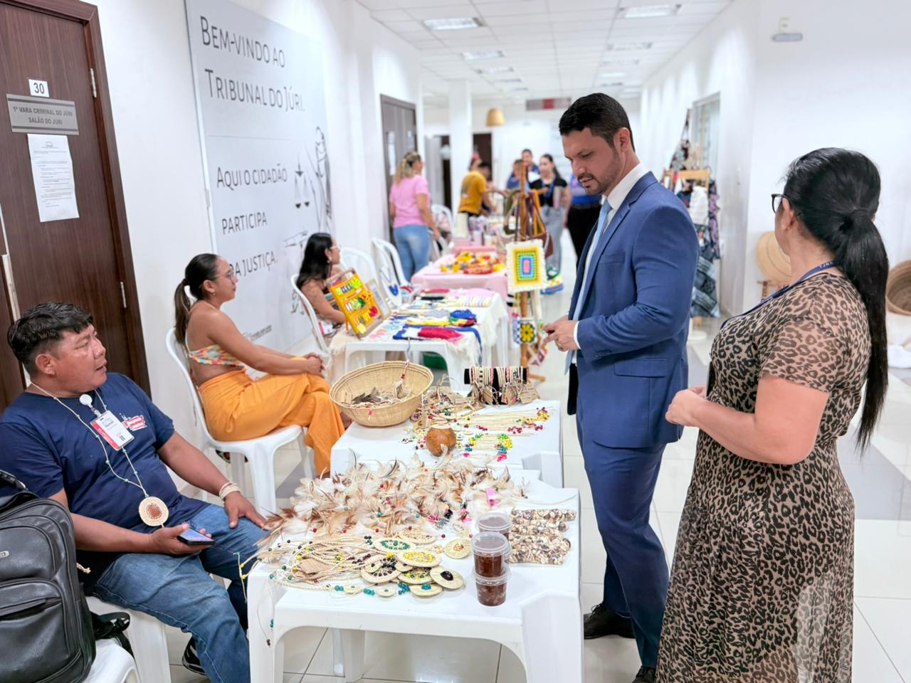 Uma foto mais ampla mostrando o corredor da feira e seu contexto. O juiz Cleber Gonçalves Filho, de terno azul e uma mulher de vestido estampado param ao lado de uma mesa branca cheia de arte com penas. Um homem indígena sentado está à esquerda da mesa. Ao fundo, há uma grande parede com texto de boas vindas ao Tribunal do Júri e uma porta com a numeração 30, ao lado de mais barracas e pessoas ao longo do corredor.