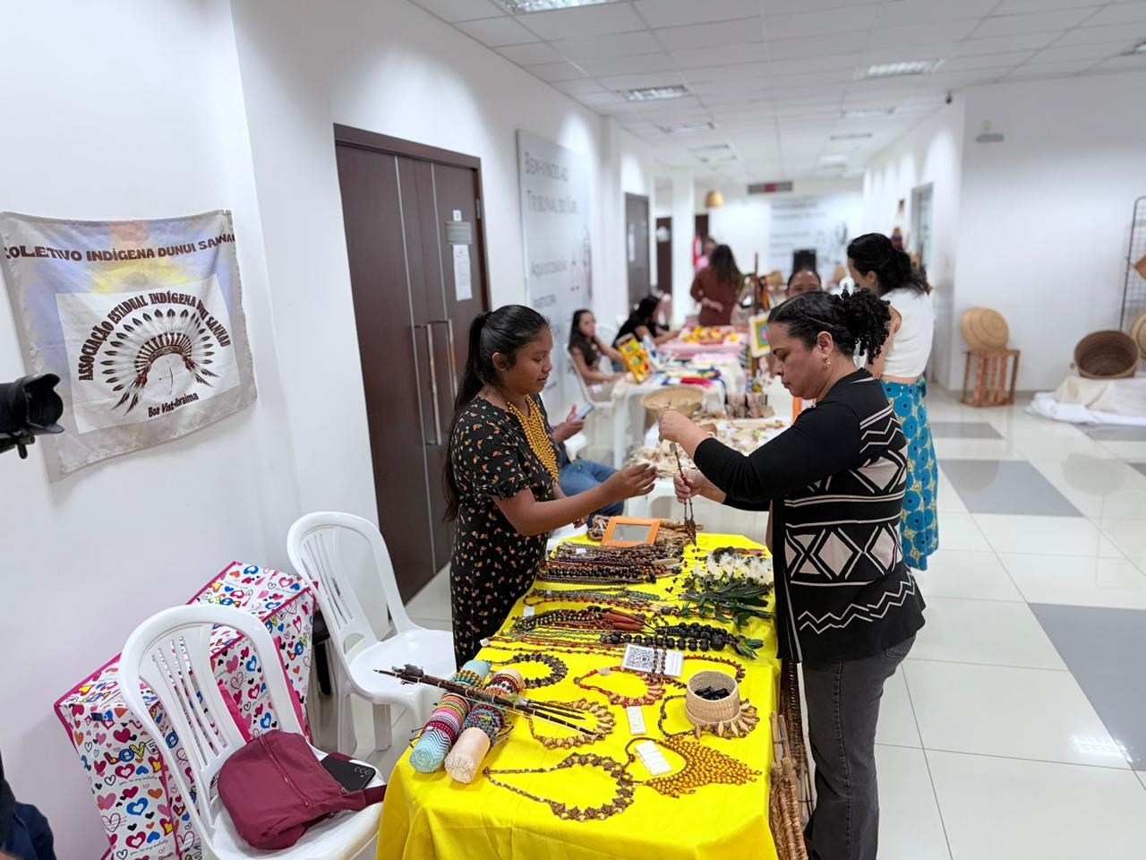  Uma foto de uma feira de artesanato indígena em um corredor interno. Uma jovem mulher indígena, com colares tradicionais, mostra um pequeno item tecido para uma mulher, em frente a uma mesa coberta com um pano amarelo repleta de biojoias, colares e acessórios tradicionais. Há cadeiras plásticas vazias ao lado e uma placa de coletivo indígena na parede à esquerda. O corredor se estende ao fundo com outras barracas e pessoas.