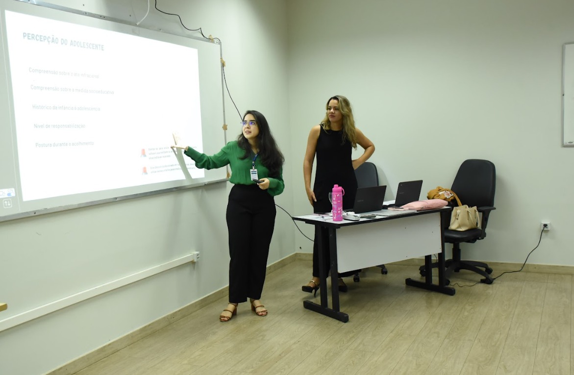 Duas mulheres em uma apresentação, com uma delas apontando para um slide com informações sobre a percepção do adolescente. Há uma mesa com laptops e objetos pessoais ao fundo.