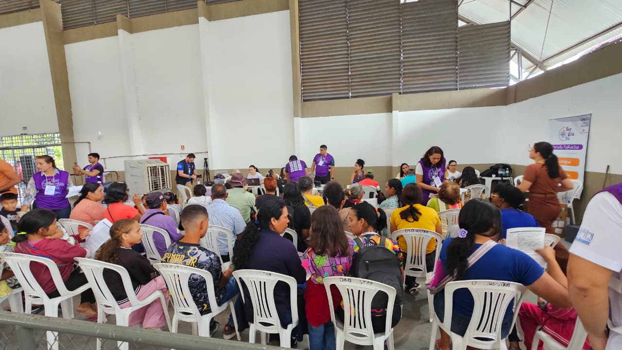 Imagem colorida mostra uma visão panorâmica de um ginásio ou galpão com várias fileiras de cadeiras brancas ocupadas por pessoas aguardando atendimento. Ao fundo e nas laterais, voluntários e profissionais com coletes roxos circulam entre o público e trabalham em mesas com laptops, organizando o fluxo de assistência durante o mutirão de serviços sociais e jurídicos.