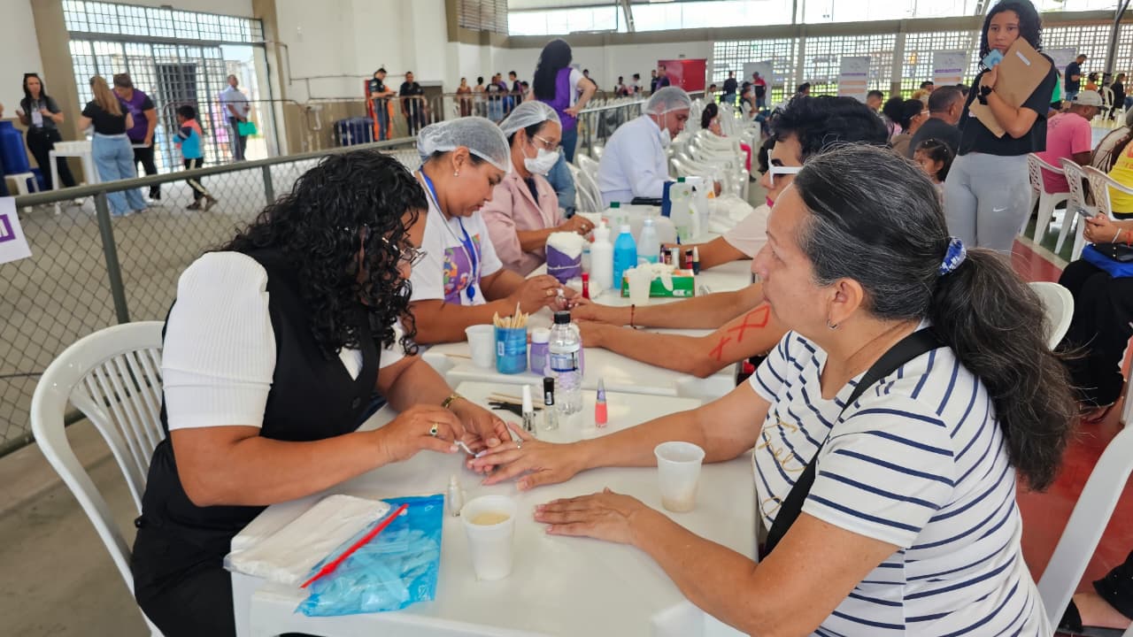 Imagem colorida mostra uma área de cuidados pessoais onde várias mulheres estão sentadas em mesas brancas recebendo serviços de manicure. As profissionais utilizam toucas e máscaras descartáveis, manuseando esmaltes e instrumentos enquanto interagem.
