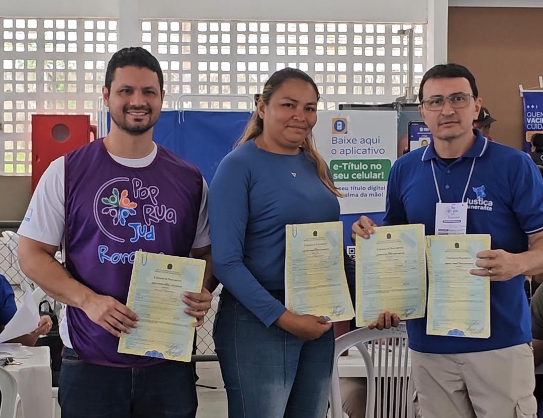 Imagem colorida mostra o juiz Cleber Gonçalves, à esquerda, vestindo um colete roxo do programa Pop Rua Jud Roraima, ao lado de uma mulher de azul e de um homem com camisa polo azul da Justiça Itinerante. Os três estão em pé, sorrindo para a câmera e segurando quatro certidões de nascimento em papel amarelado, em um ambiente amplo e iluminado.