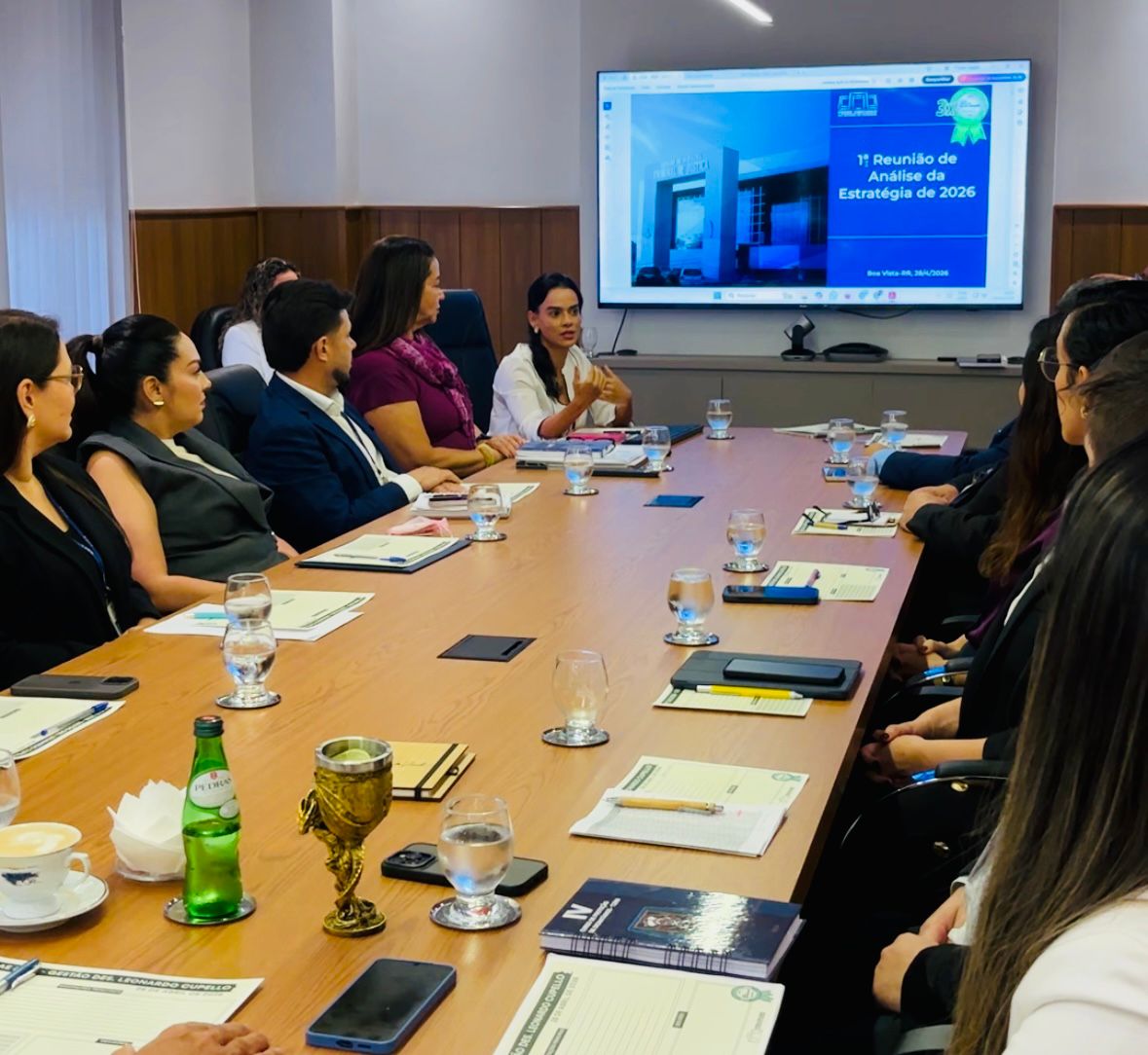 Imagem em plano aberto de  secretários e coordenadores reunidos durante reunião de alinhamento. Ao fundo é possível ver uma TV com com apresentação da temática. 