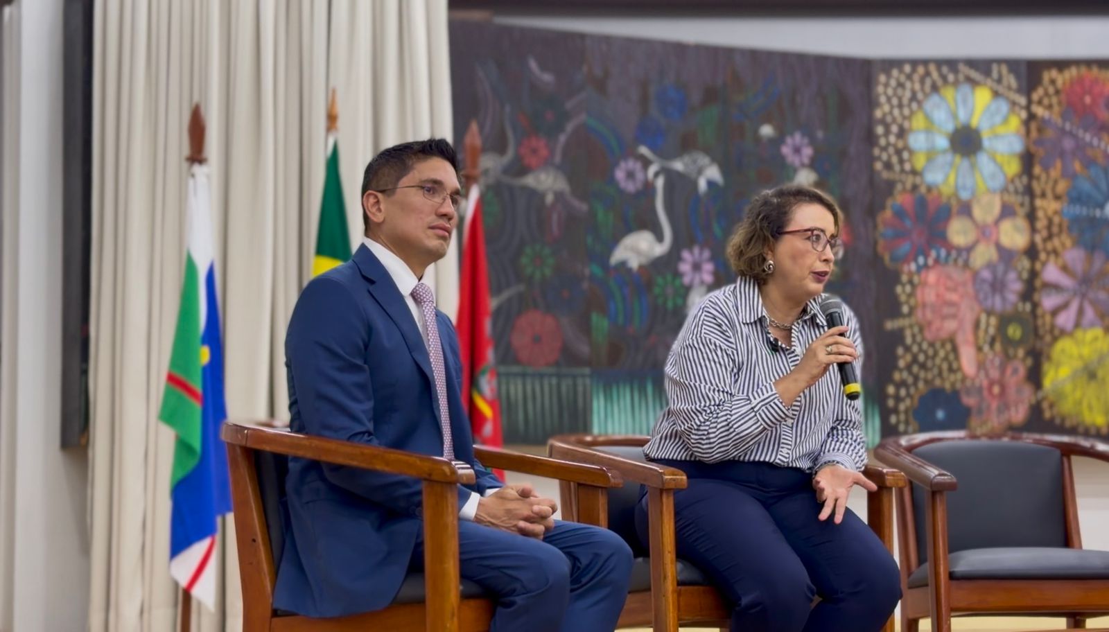 Em um palco, a juíza Dra. Lana Leitão e o juiz Dr. Marcelo Oliveira estão sentados em poltronas de madeira. A juíza fala ao microfone enquanto o juiz a acompanha. Ao fundo, há cortinas claras, bandeiras oficiais e um grande painel artístico colorido com flores e aves.