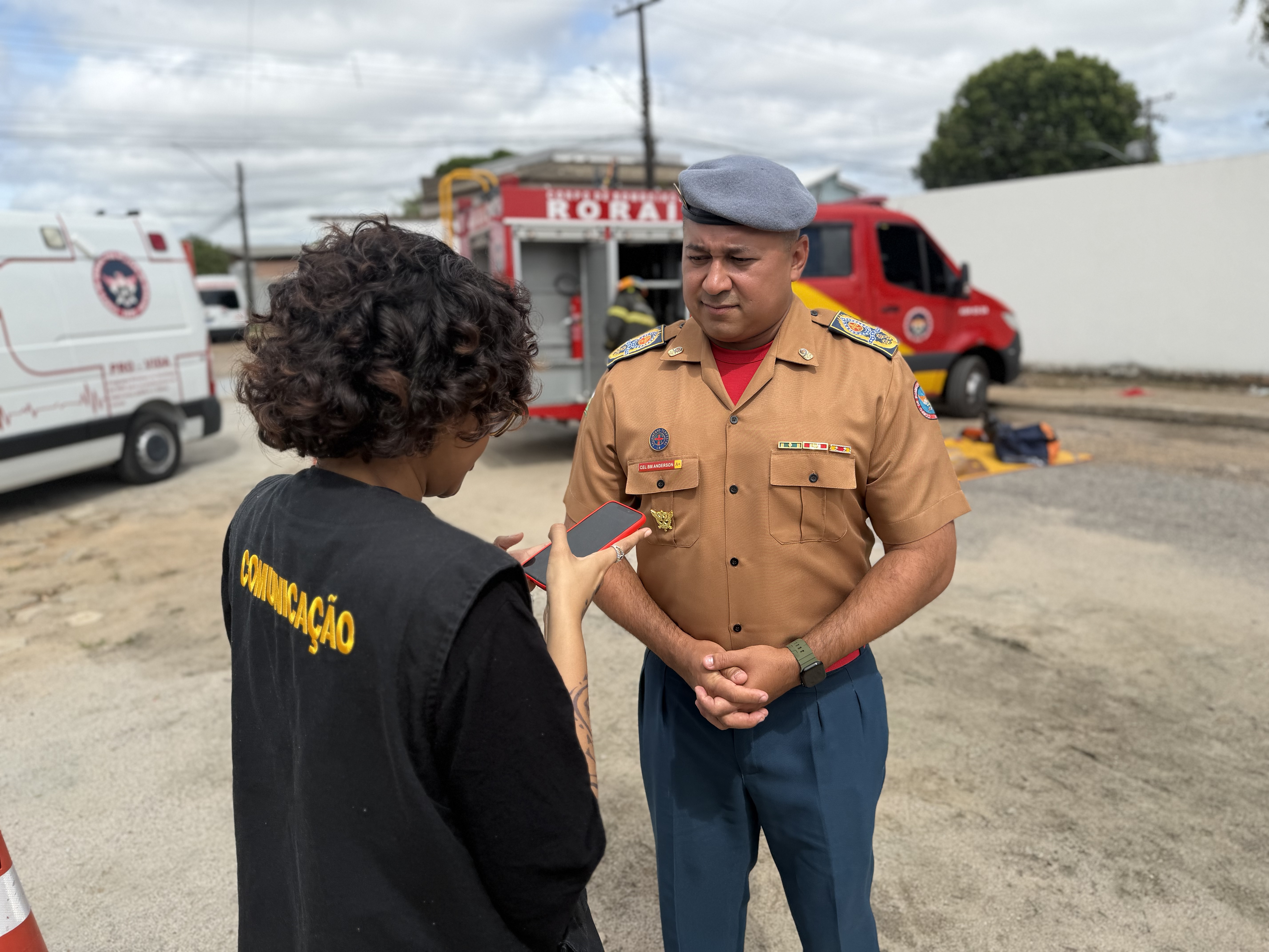 O Coronel Anderson Matos, fardado, concede uma entrevista para uma profissional da equipe de comunicação, que segura um celular para gravar o áudio. Ela veste um colete preto com a palavra “COMUNICAÇÃO” escrita em amarelo nas costas. O Coronel Anderson Matos, fardado, concede uma entrevista para uma profissional da equipe de comunicação, que segura um celular para gravar o áudio. Ela veste um colete preto com a palavra “COMUNICAÇÃO” escrita em amarelo nas costas.