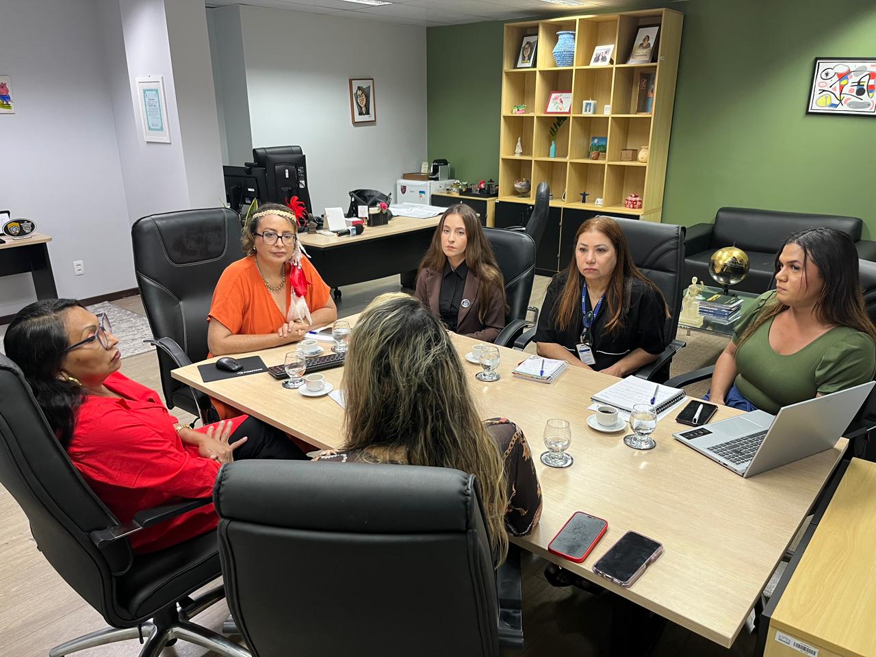 Imagem colorida mostra um grupo de seis mulheres reunidas em torno de uma mesa de reunião clara, conversando em um ambiente de escritório com notebooks, xícaras de café e uma estante de livros ao fundo.