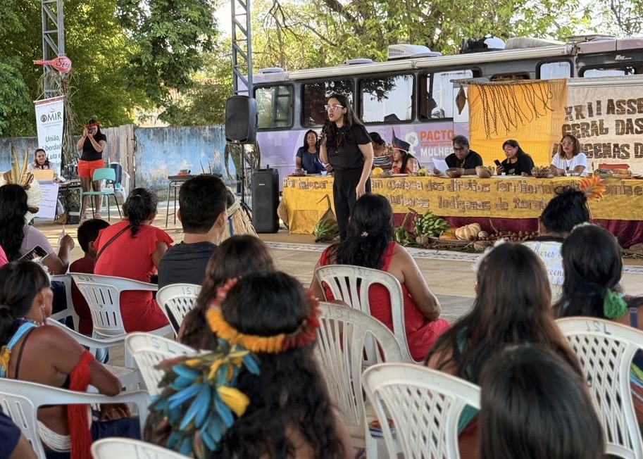  A juíza Dra. Suelen Alves fala ao microfone para uma plateia composta majoritariamente por mulheres indígenas, sentadas em cadeiras brancas em um espaço ao ar livre. Ao fundo, há uma mesa comprida com pessoas sentadas, e atrás dela um ônibus com inscrições.