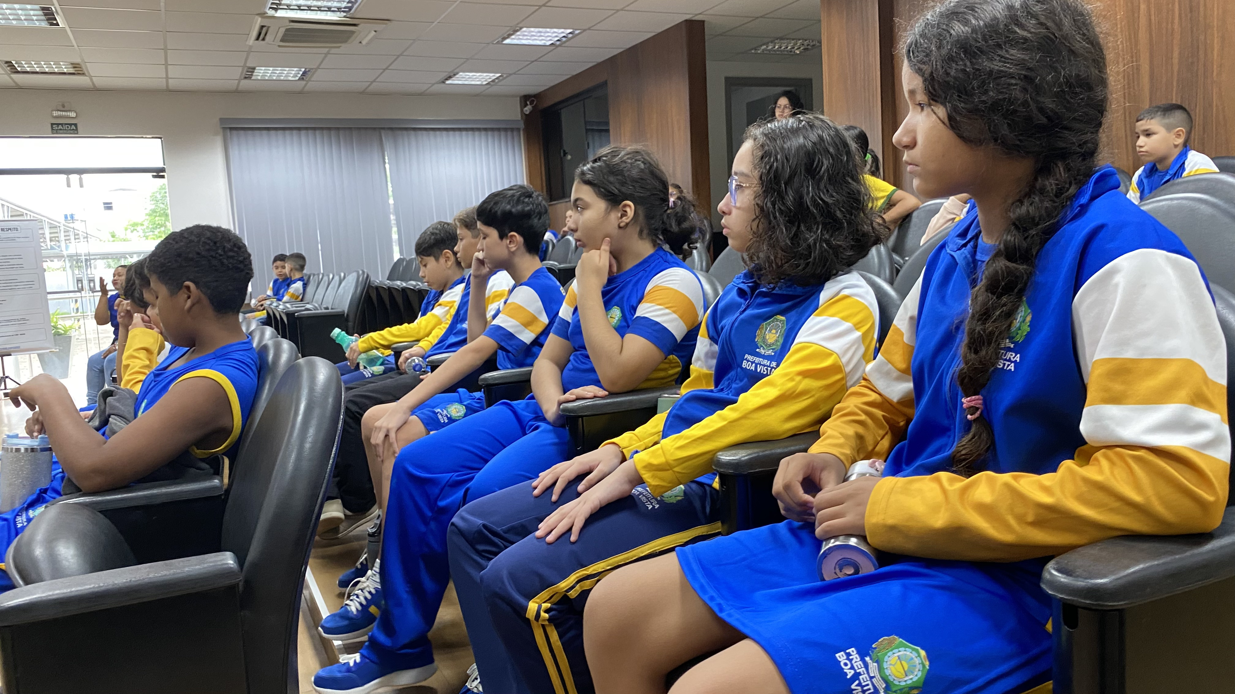  Uma fileira de estudantes vestindo uniformes azuis e amarelos da Prefeitura de Boa Vista sentados em poltronas pretas de um auditório, assistindo atentamente a uma atividade à frente.