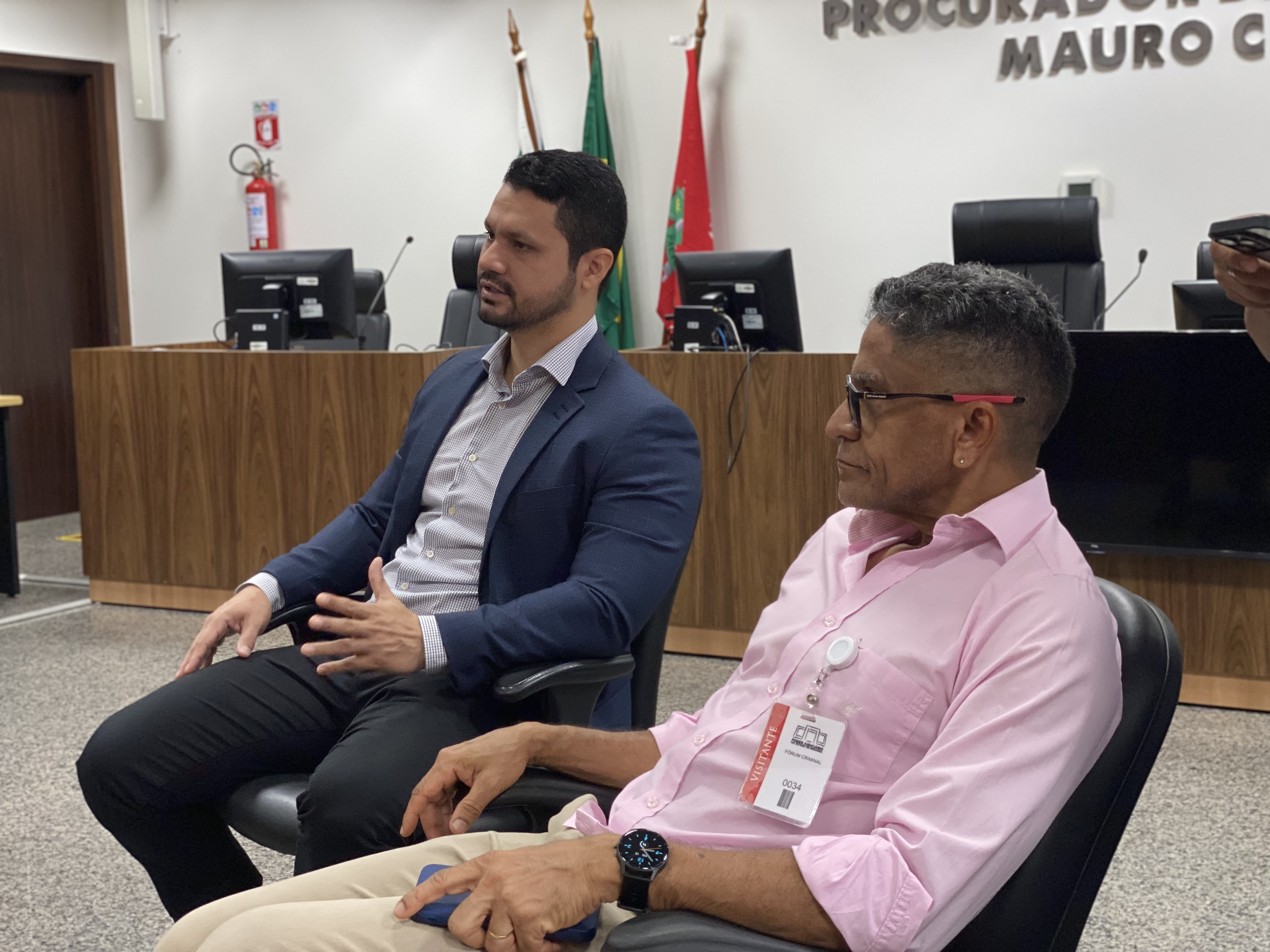  Juiz Cleber Gonçalves e um dos professores da turma sentados de perfil em cadeiras de escritório. O juiz à esquerda veste paletó escuro sobre camisa clara e gesticula enquanto fala; o professor à direita veste camisa rosa, óculos e usa um crachá de visitante. Ao fundo, vê-se a bancada de um plenário com computadores e bandeiras oficiais.