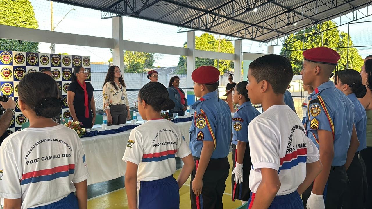  Cerimônia com alunos de fardamento militar, em pé, acompanhando falas de autoridades em ambiente interno decorado com banners.