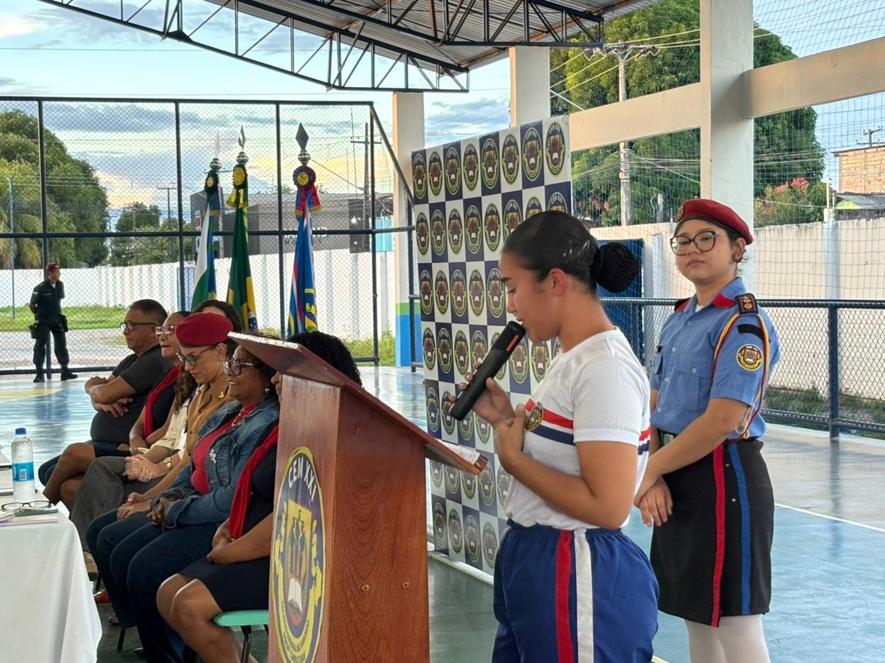 Menina em uniforme fala ao microfone em evento, enquanto outras pessoas assistem sentadas. Bandeiras ao fundo e duas mulheres em uniforme de segurança.