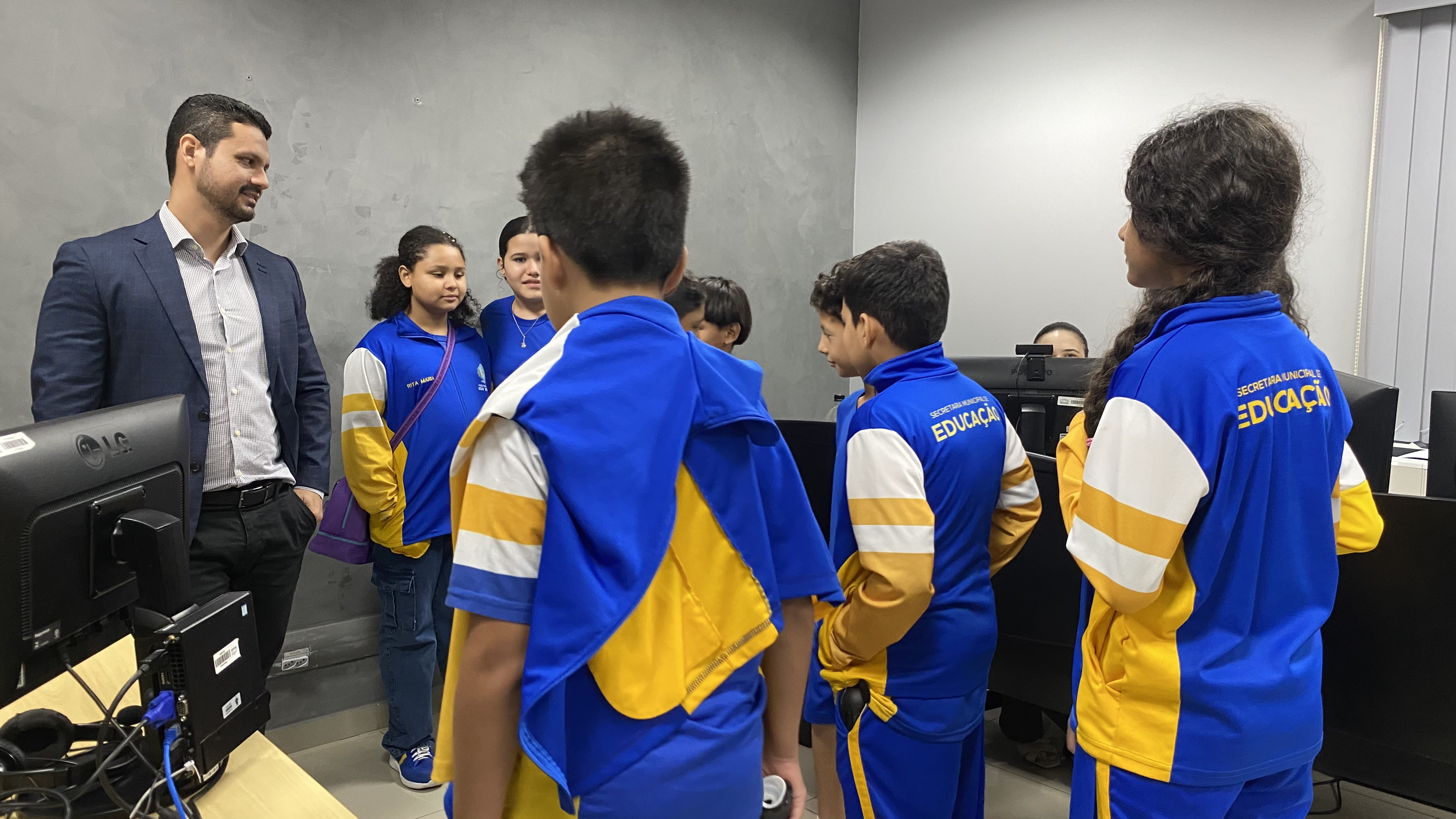O juiz de paletó escuro está de pé à esquerda, sorrindo e conversando com um grupo de estudantes em uma sala de escritório. Os alunos vestem o uniforme azul e amarelo e estão reunidos entre bancadas com computadores e monitores. Ao fundo, a parede é cinza escuro texturizada.