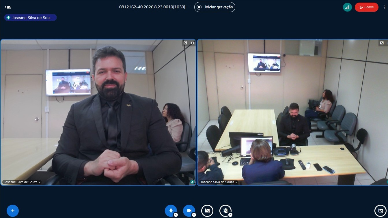 Captura de tela colorida mostra uma audiência por videoconferência. A tela está dividida em duas janelas: à esquerda, aparece em destaque um intérprete de Libras usando terno preto e fazendo sinais com as mãos; à direita, a sala da audiência é vista em plano aberto, com pessoas sentadas à mesa, equipamentos de videoconferência e uma tela fixada na parede exibindo a reunião virtual.