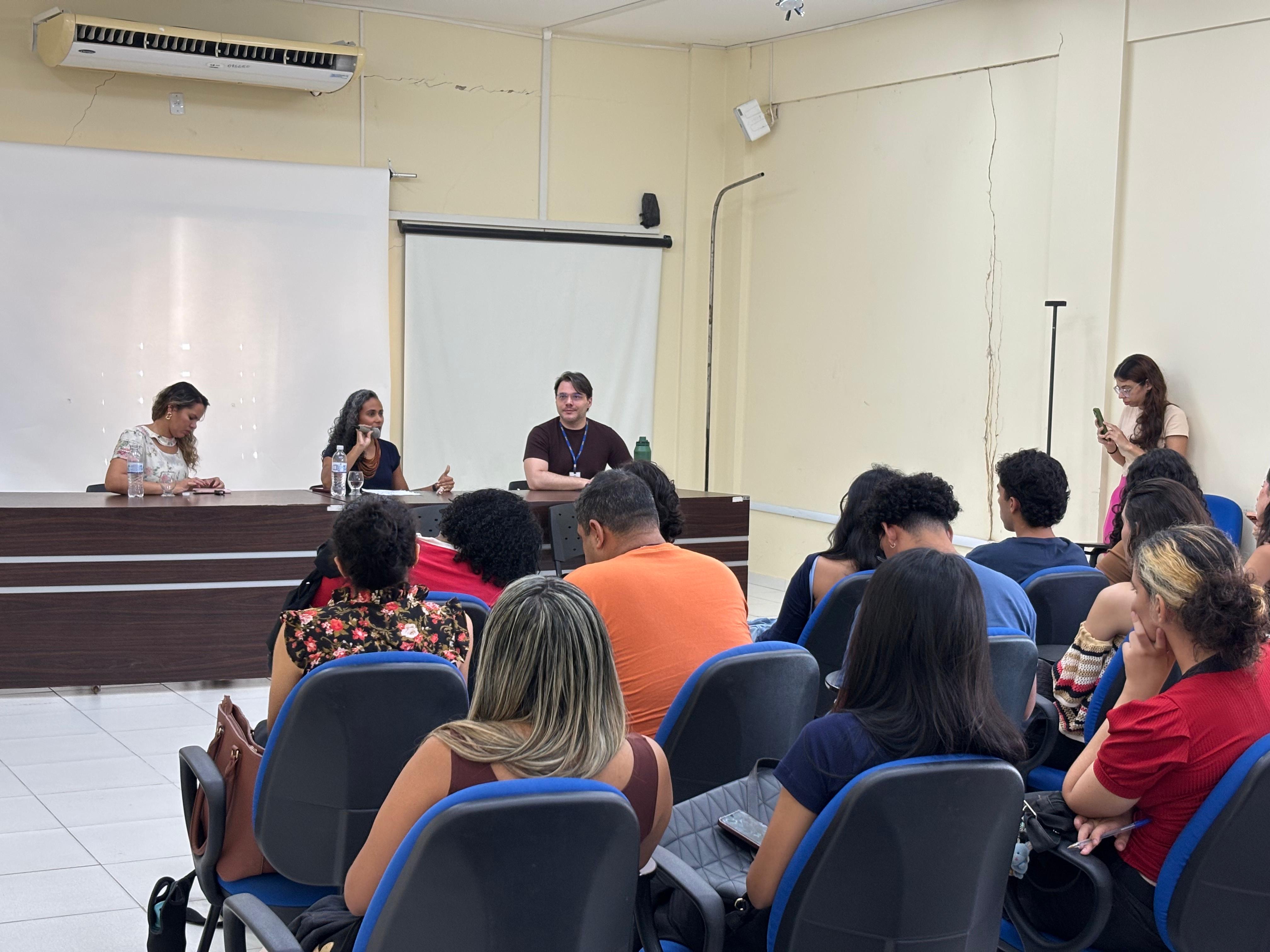 imagem colorida em um auditório interno é possível ver acadêmicos sentados. Na frente há uma mesa de composta de três pessoas, sendo que a mulher do é servidora do TJRR e está falando ao microfone. imagem colorida em um auditório interno é possível ver acadêmicos sentados. Na frente há uma mesa de composta de três pessoas, sendo que a mulher do é servidora do TJRR e está falando ao microfone.