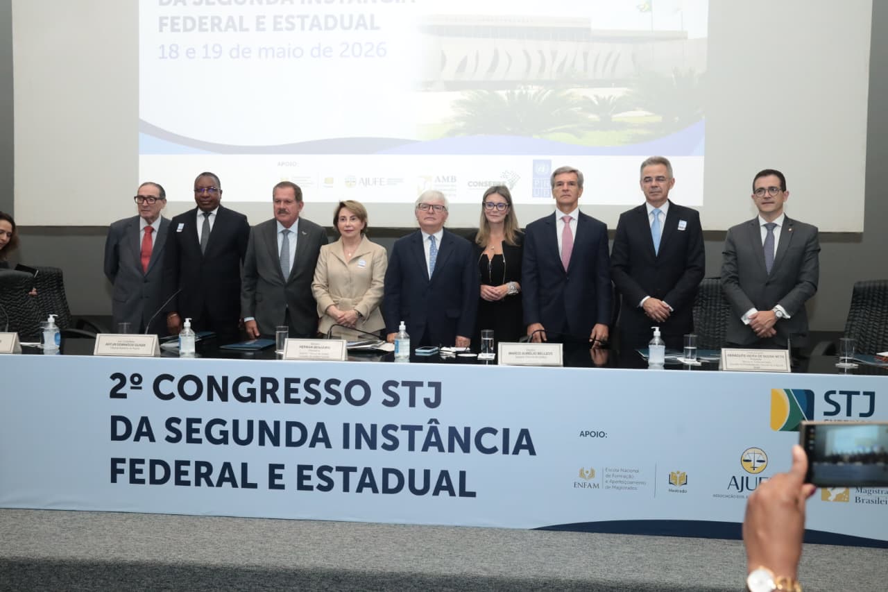 Nove autoridades (sete homens e duas mulheres) vestindo trajes formais estão de pé, alinhadas atrás de uma longa bancada azul. Na frente da bancada, destaca-se o título em letras escuras: "2º CONGRESSO STJ DA SEGUNDA INSTÂNCIA FEDERAL E ESTADUAL". Ao fundo, um grande telão projeta o mesmo texto do evento. No canto inferior direito, uma mão segura um celular registrando o momento. Nove autoridades (sete homens e duas mulheres) vestindo trajes formais estão de pé, alinhadas atrás de uma longa bancada azul. Na frente da bancada, destaca-se o título em letras escuras: "2º CONGRESSO STJ DA SEGUNDA INSTÂNCIA FEDERAL E ESTADUAL". Ao fundo, um grande telão projeta o mesmo texto do evento. No canto inferior direito, uma mão segura um celular registrando o momento.
