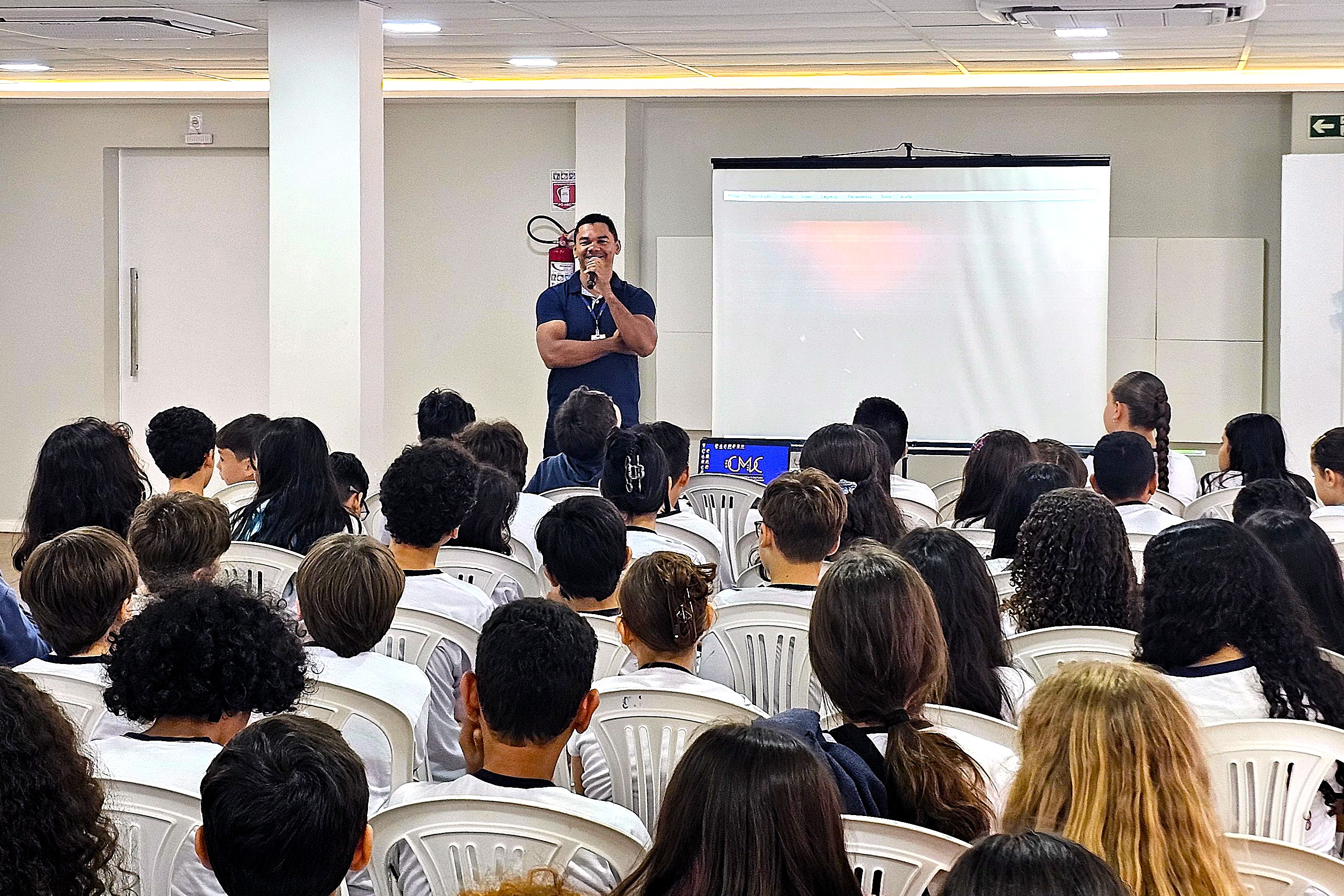 Foto colorida mostra estudantes sentados em cadeiras brancas durante uma apresentação no Centro de Memória e Cultura. À frente, um servidor fala ao microfone próximo a uma tela de projeção, enquanto o público acompanha a atividade. Foto colorida mostra estudantes sentados em cadeiras brancas durante uma apresentação no Centro de Memória e Cultura. À frente, um servidor fala ao microfone próximo a uma tela de projeção, enquanto o público acompanha a atividade.