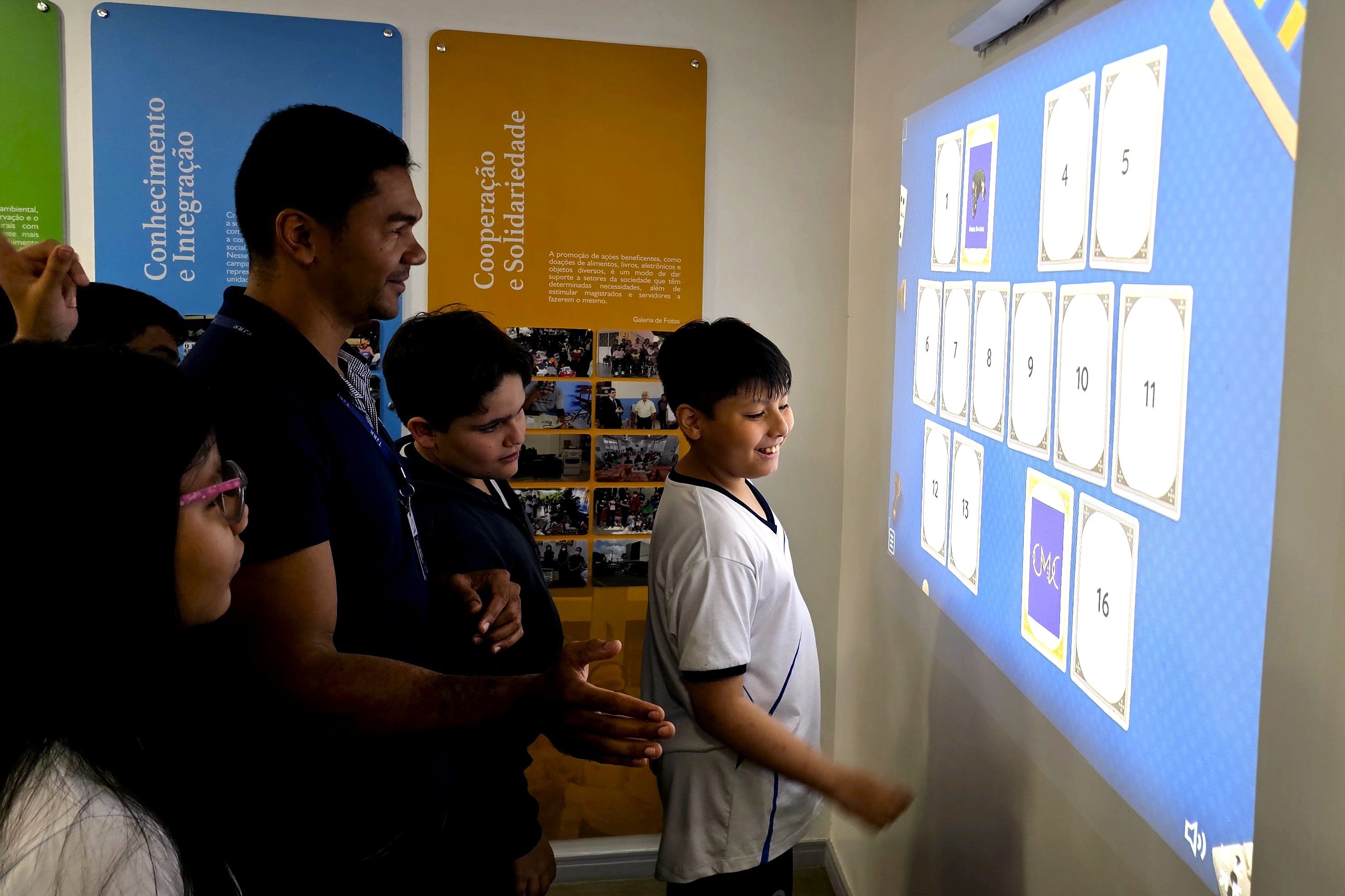 Foto colorida mostra estudantes participando de uma atividade interativa com projeção na parede. Um menino interage com um jogo de cartas exibido na tela, enquanto outros estudantes e um adulto acompanham a dinâmica no Centro de Memória e Cultura. Foto colorida mostra estudantes participando de uma atividade interativa com projeção na parede. Um menino interage com um jogo de cartas exibido na tela, enquanto outros estudantes e um adulto acompanham a dinâmica no Centro de Memória e Cultura.