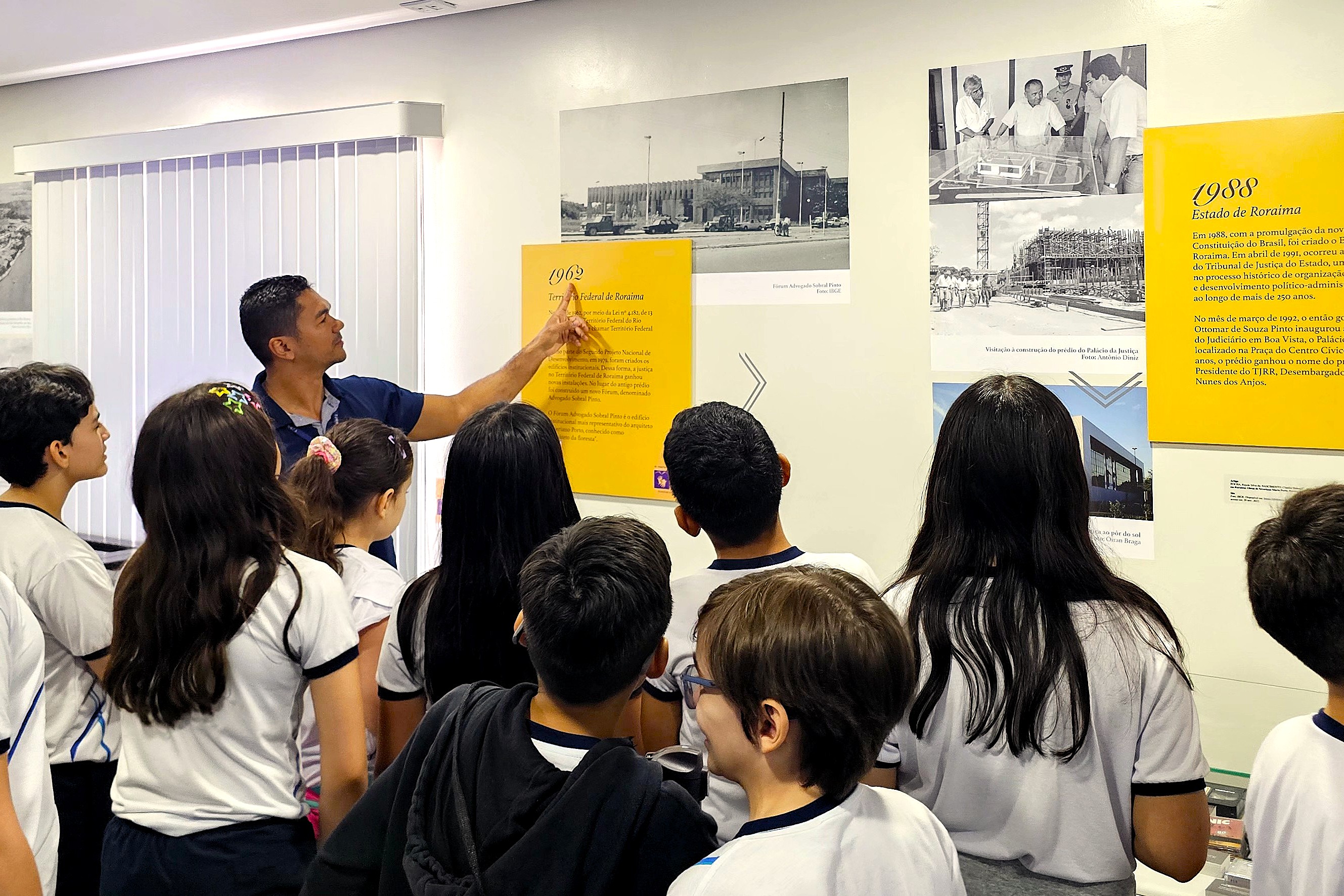 Foto colorida mostra estudantes em visita guiada a uma exposição histórica. Um servidor aponta para um painel na parede, enquanto o grupo observa fotografias antigas e textos sobre a história de Roraima e do Poder Judiciário. Foto colorida mostra estudantes em visita guiada a uma exposição histórica. Um servidor aponta para um painel na parede, enquanto o grupo observa fotografias antigas e textos sobre a história de Roraima e do Poder Judiciário.