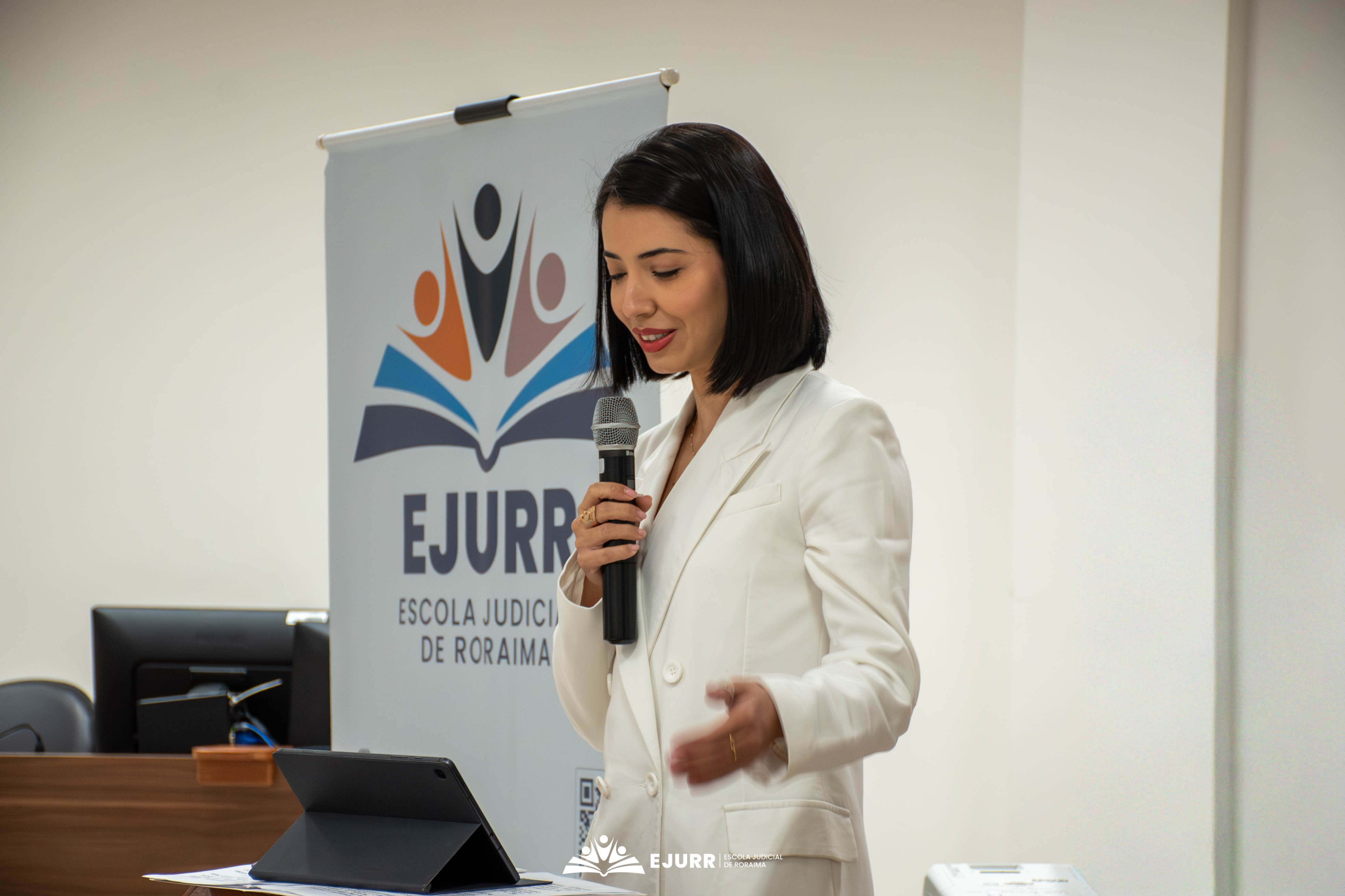 Uma palestrante de cabelos curtos escuros e blazer branco fala ao microfone, posicionada ao lado de uma bancada com um tablet. Atrás dela, há um banner institucional cinza com o logotipo colorido da EJURR. Uma palestrante de cabelos curtos escuros e blazer branco fala ao microfone, posicionada ao lado de uma bancada com um tablet. Atrás dela, há um banner institucional cinza com o logotipo colorido da EJURR.