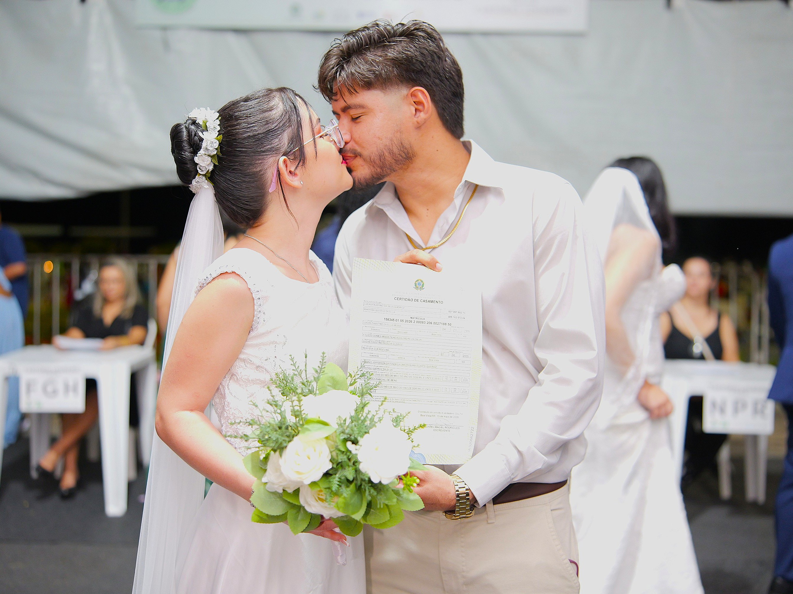 Foto colorida mostra um casal de noivos se beijando após a oficialização da união. A noiva usa vestido branco e segura um buquê, enquanto o noivo segura a certidão de casamento. Ao fundo, outras pessoas aparecem no espaço da cerimônia. Foto colorida mostra um casal de noivos se beijando após a oficialização da união. A noiva usa vestido branco e segura um buquê, enquanto o noivo segura a certidão de casamento. Ao fundo, outras pessoas aparecem no espaço da cerimônia.