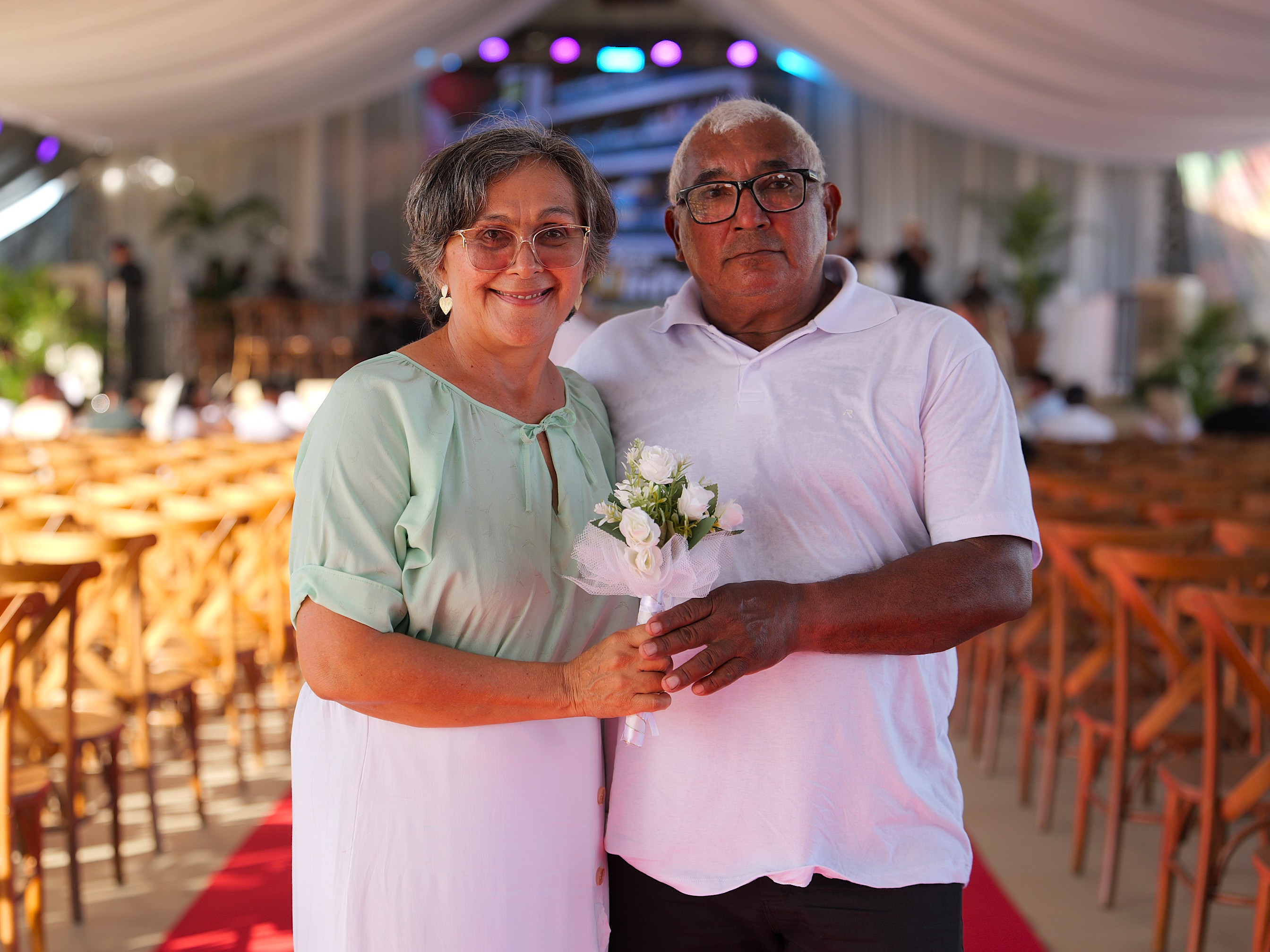 Foto colorida mostra um casal posando em um espaço decorado para cerimônia. A mulher segura um pequeno buquê de flores brancas, enquanto o homem está ao lado dela. Ao fundo, aparecem cadeiras e iluminação do ambiente. Foto colorida mostra um casal posando em um espaço decorado para cerimônia. A mulher segura um pequeno buquê de flores brancas, enquanto o homem está ao lado dela. Ao fundo, aparecem cadeiras e iluminação do ambiente.