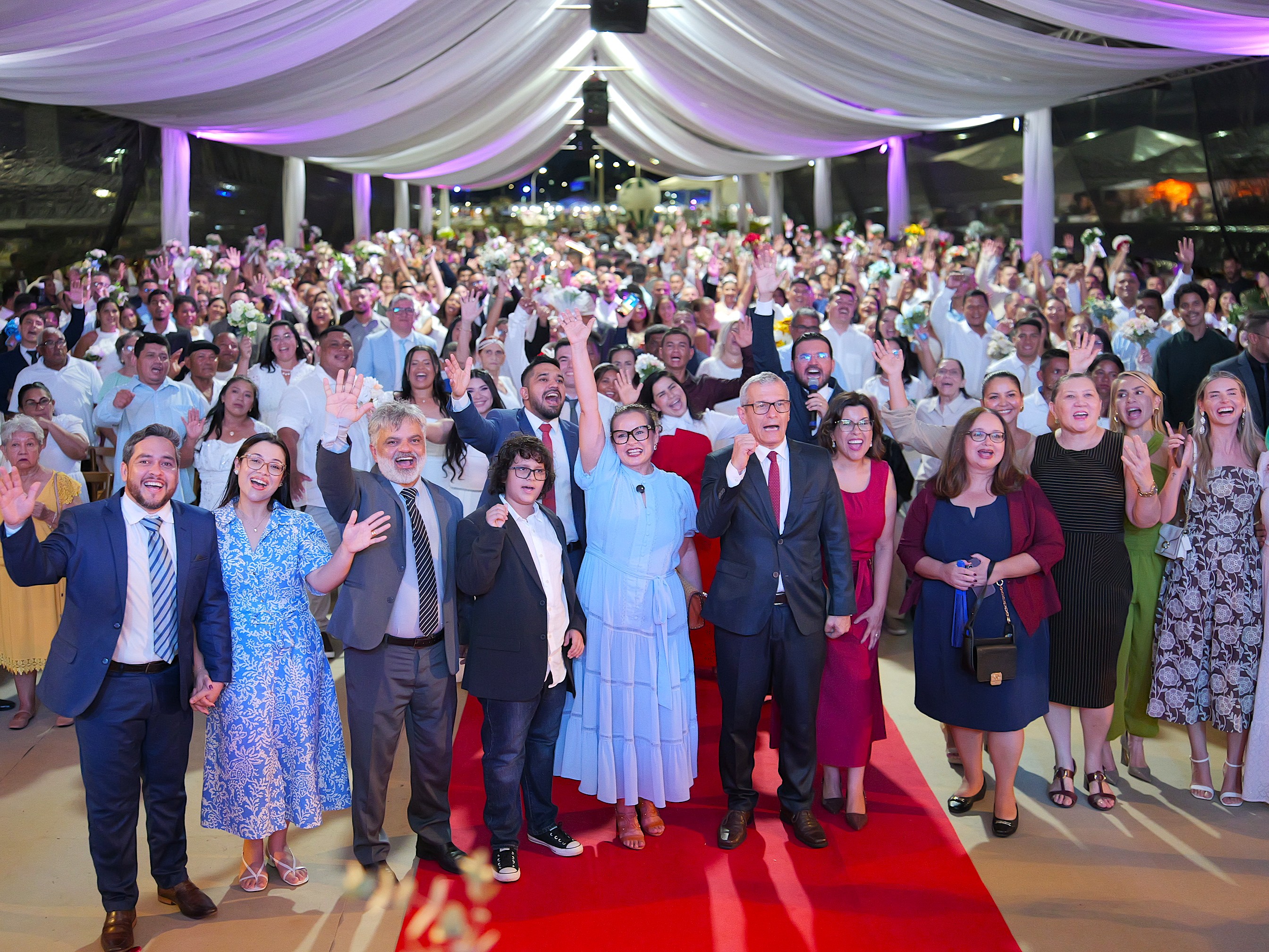 Foto colorida mostra uma grande quantidade de casais, familiares, convidados e autoridades reunidos durante o casamento coletivo. Muitas pessoas sorriem e acenam para a câmera, em um ambiente decorado com tecido branco e iluminação colorida. Foto colorida mostra uma grande quantidade de casais, familiares, convidados e autoridades reunidos durante o casamento coletivo. Muitas pessoas sorriem e acenam para a câmera, em um ambiente decorado com tecido branco e iluminação colorida.