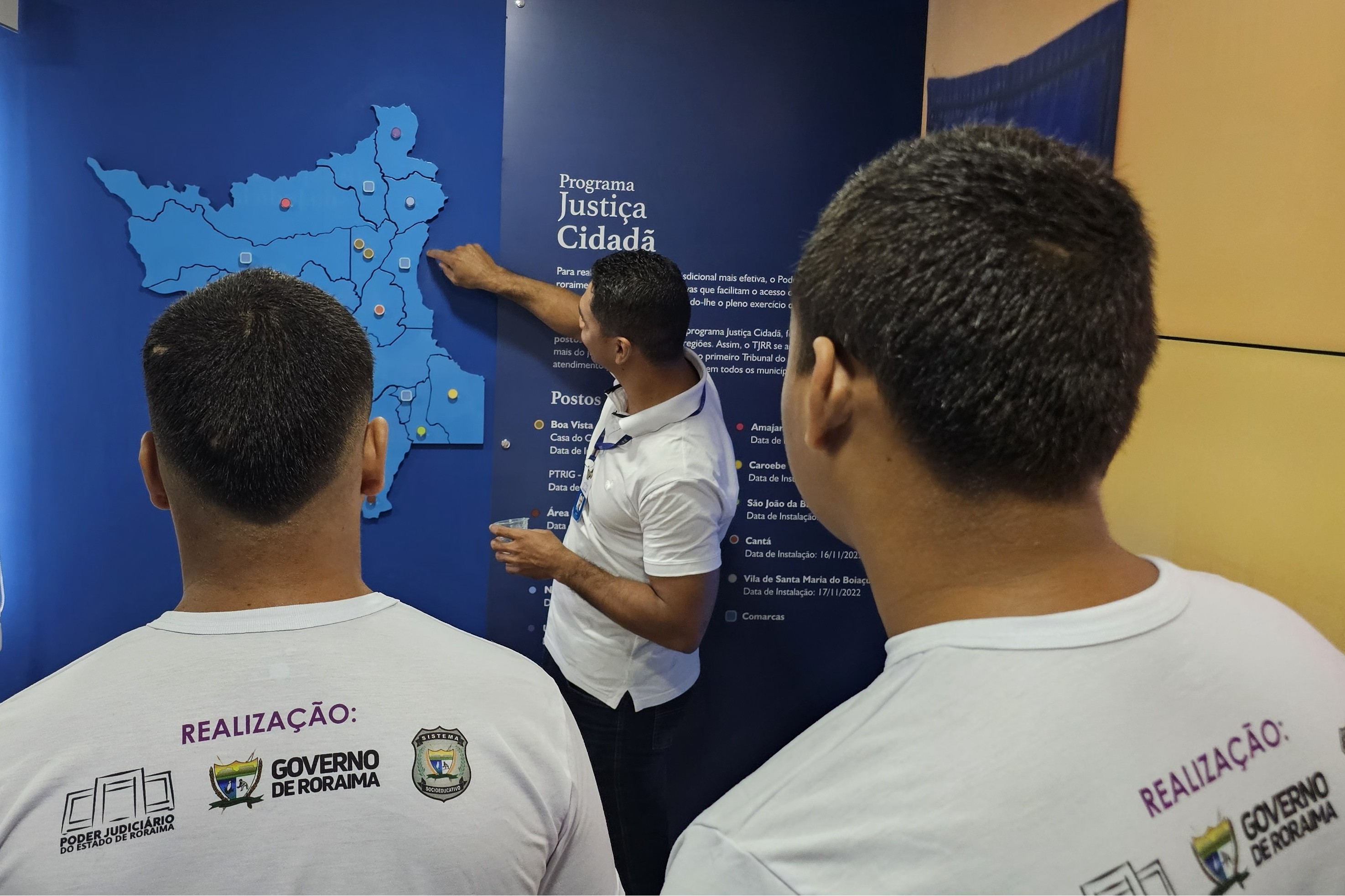 Foto colorida mostra jovens observando um painel sobre o Programa Justiça Cidadã durante visita guiada. Um servidor aponta para um mapa de Roraima fixado na parede, indicando pontos de atendimento no estado. Os participantes aparecem de costas, acompanhando a explicação. Foto colorida mostra jovens observando um painel sobre o Programa Justiça Cidadã durante visita guiada. Um servidor aponta para um mapa de Roraima fixado na parede, indicando pontos de atendimento no estado. Os participantes aparecem de costas, acompanhando a explicação.