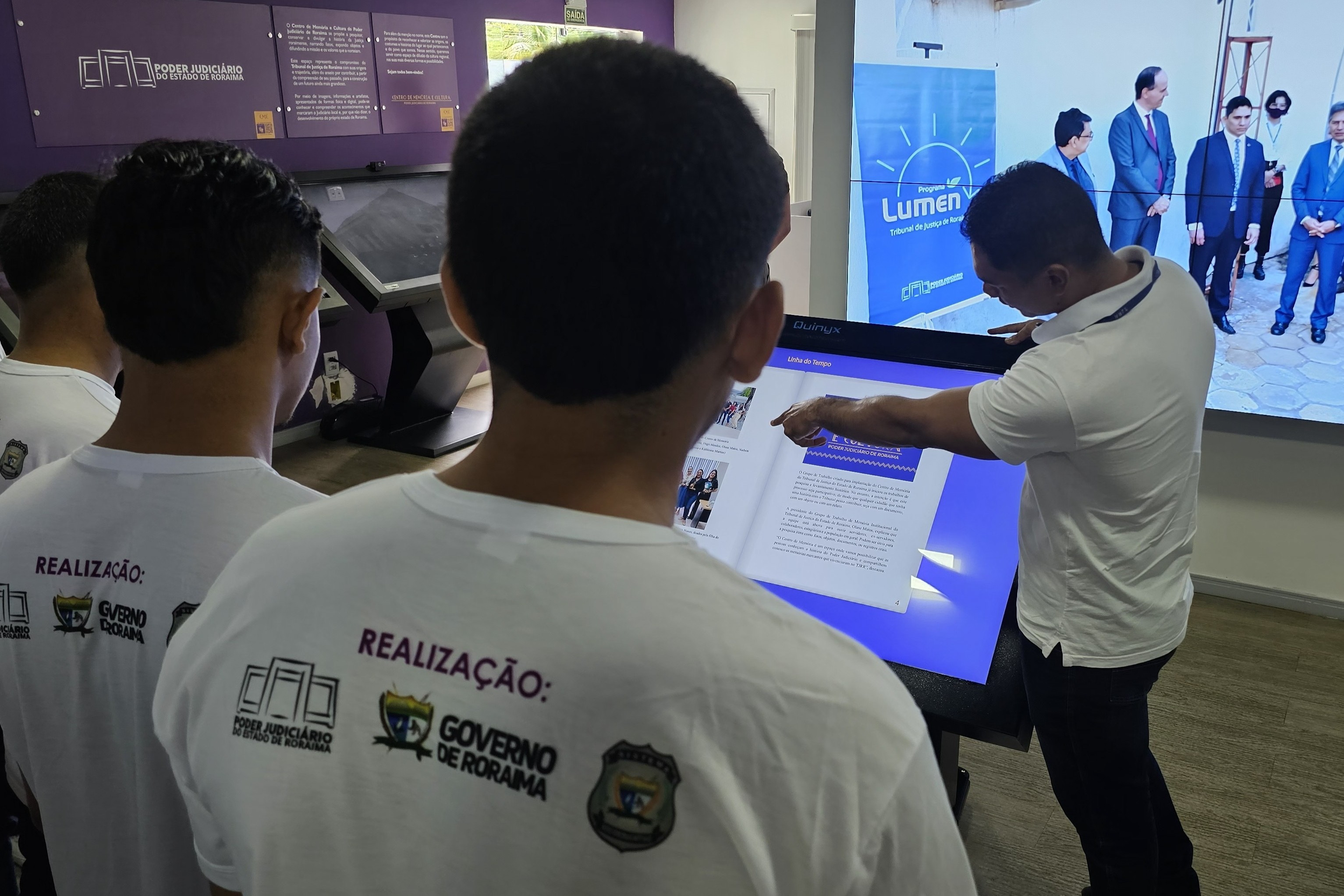 Foto colorida mostra jovens acompanhando uma explicação durante visita ao Centro de Memória e Cultura do Judiciário. Um servidor aponta para uma tela interativa com conteúdo sobre a linha do tempo do TJRR. Ao fundo, uma tela maior exibe imagem relacionada ao Programa Lúmen. Foto colorida mostra jovens acompanhando uma explicação durante visita ao Centro de Memória e Cultura do Judiciário. Um servidor aponta para uma tela interativa com conteúdo sobre a linha do tempo do TJRR. Ao fundo, uma tela maior exibe imagem relacionada ao Programa Lúmen.