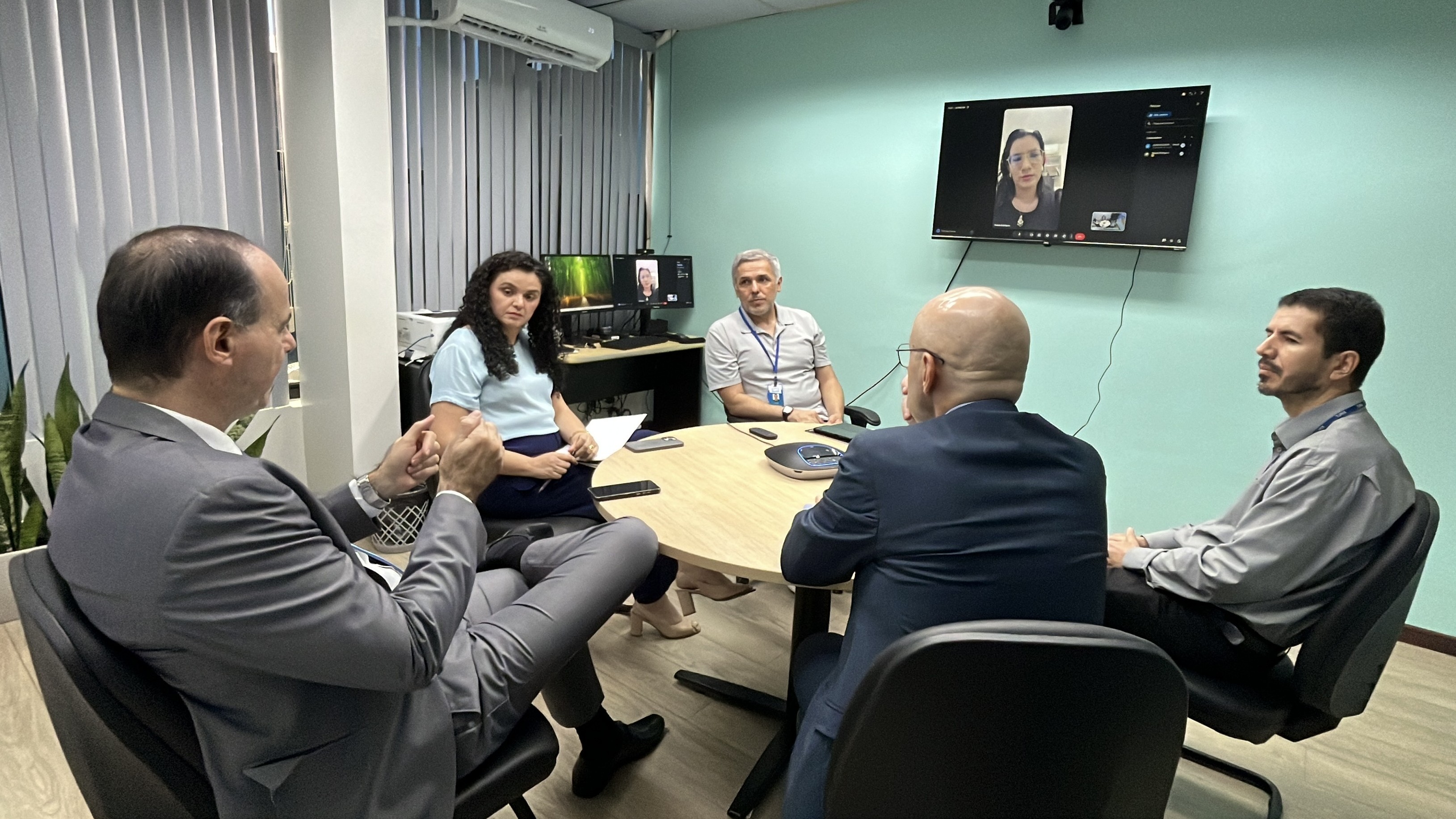 Foto colorida mostra reunião híbrida em uma sala do Tribunal de Justiça de Roraima. Participantes estão sentados ao redor de uma mesa, enquanto uma mulher aparece por videoconferência em uma tela fixada na parede. Sobre a mesa há documentos, celulares e equipamento de áudio. Foto colorida mostra reunião híbrida em uma sala do Tribunal de Justiça de Roraima. Participantes estão sentados ao redor de uma mesa, enquanto uma mulher aparece por videoconferência em uma tela fixada na parede. Sobre a mesa há documentos, celulares e equipamento de áudio.