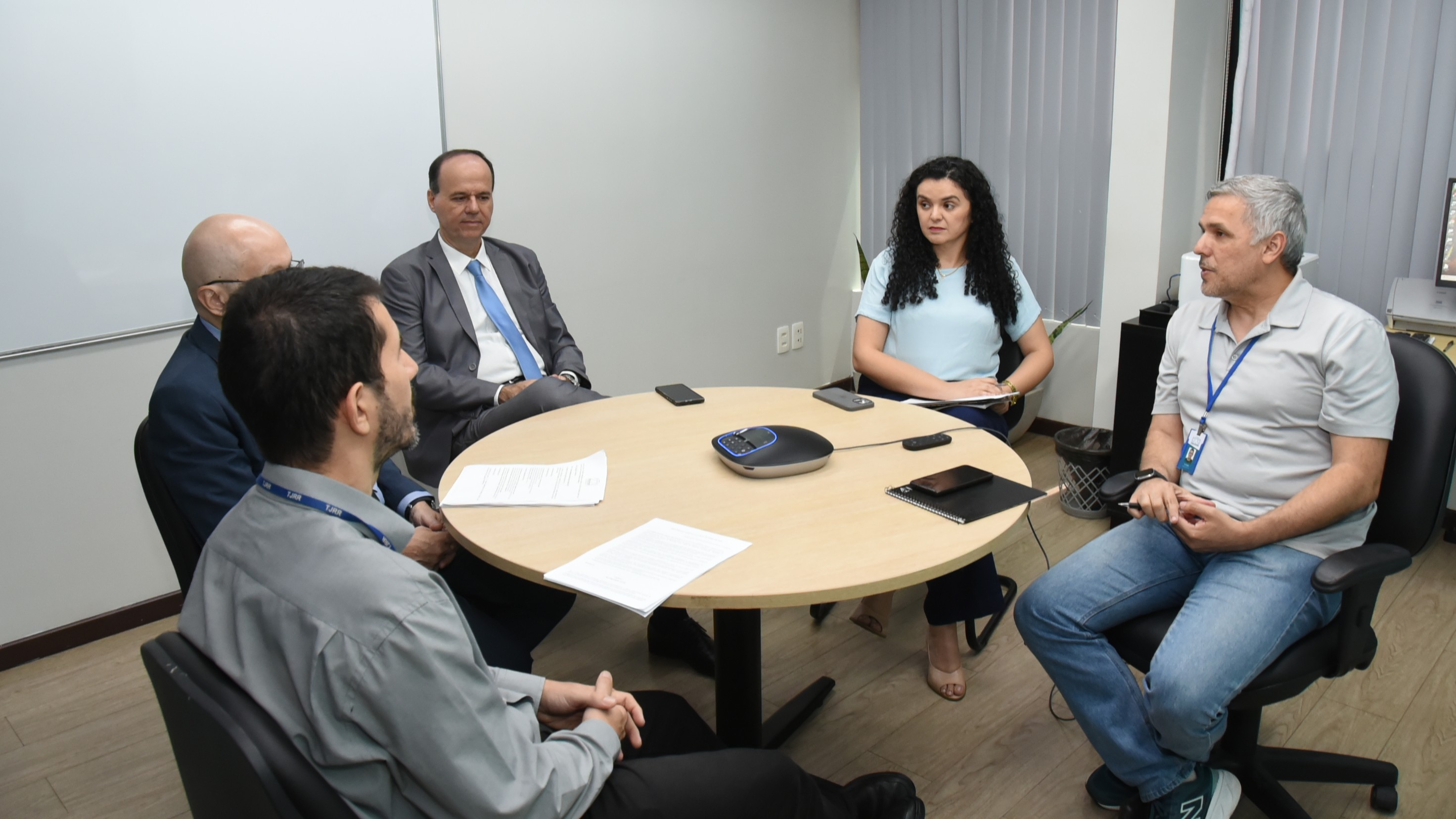 Foto colorida mostra participantes reunidos em uma sala de trabalho, sentados ao redor de uma mesa redonda. Sobre a mesa há documentos, celulares e um equipamento de áudio para videoconferência. As pessoas conversam durante encontro sobre a expansão do projeto PROVER para o posto da Justiça Cidadã no Amajari. Foto colorida mostra participantes reunidos em uma sala de trabalho, sentados ao redor de uma mesa redonda. Sobre a mesa há documentos, celulares e um equipamento de áudio para videoconferência. As pessoas conversam durante encontro sobre a expansão do projeto PROVER para o posto da Justiça Cidadã no Amajari.