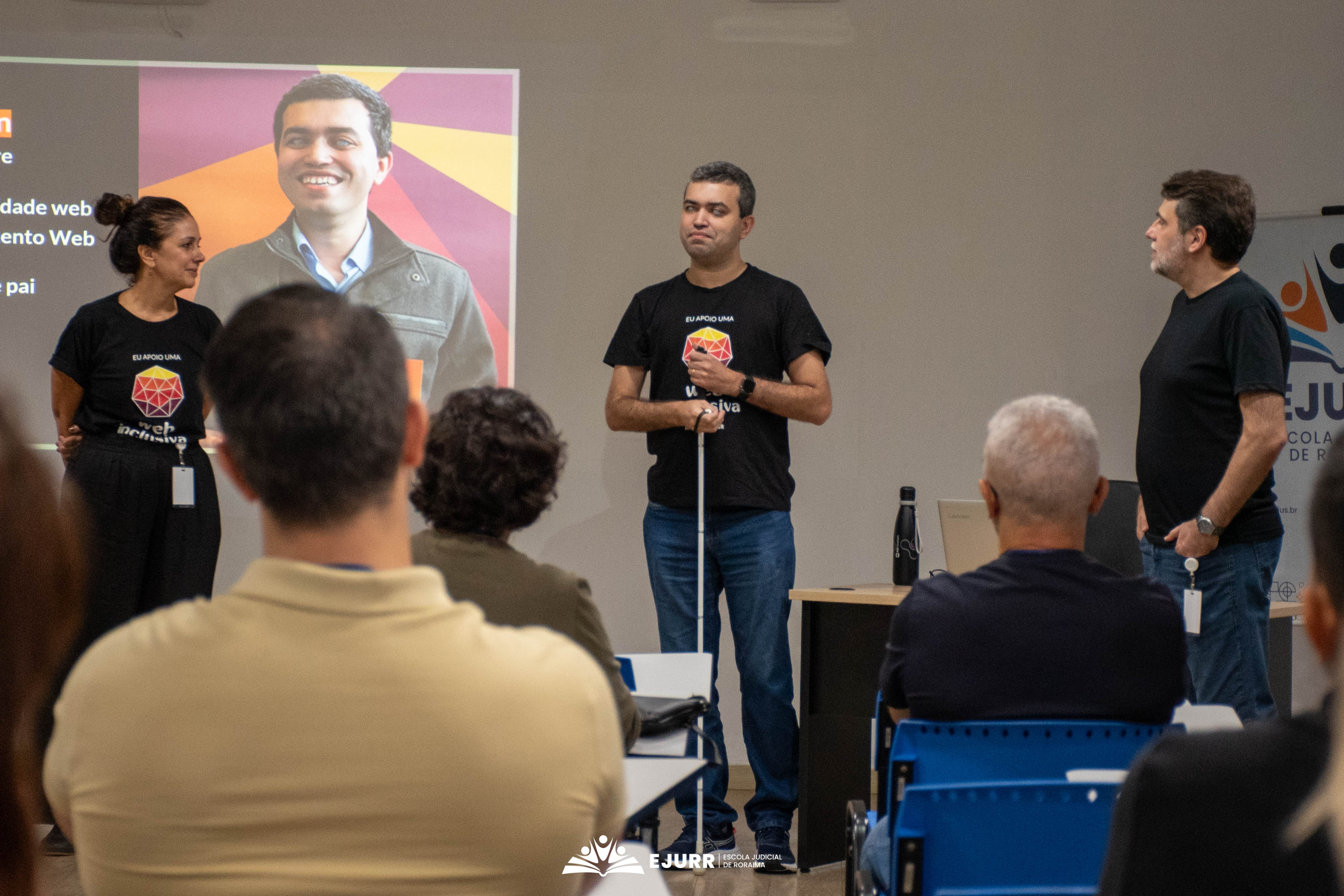 Close de três palestrantes à frente de um auditório. No centro, um homem cego veste camiseta preta escrito "Web Inclusiva" e segura uma bengala longa branca. À esquerda, uma mulher o observa, e à direita, outro homem em pé. Ao fundo, uma tela projeta a foto sorridente do palestrante cego. Em primeiro plano, as costas da cabeça de espectadores desfocados. Close de três palestrantes à frente de um auditório. No centro, um homem cego veste camiseta preta escrito "Web Inclusiva" e segura uma bengala longa branca. À esquerda, uma mulher o observa, e à direita, outro homem em pé. Ao fundo, uma tela projeta a foto sorridente do palestrante cego. Em primeiro plano, as costas da cabeça de espectadores desfocados.