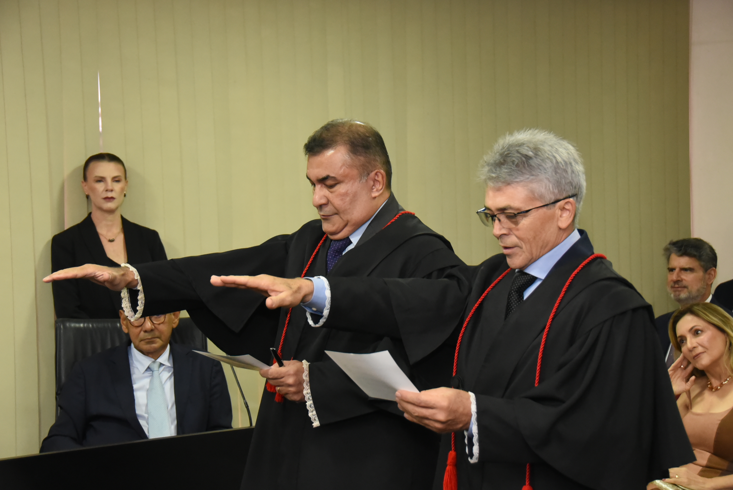 Dois procuradores, trajando vestes talares, realizam o compromisso de posse durante sessão solene. Ambos mantêm o braço direito estendido enquanto leem o termo oficial. Ao fundo, autoridades e convidados acompanham a cerimônia.
