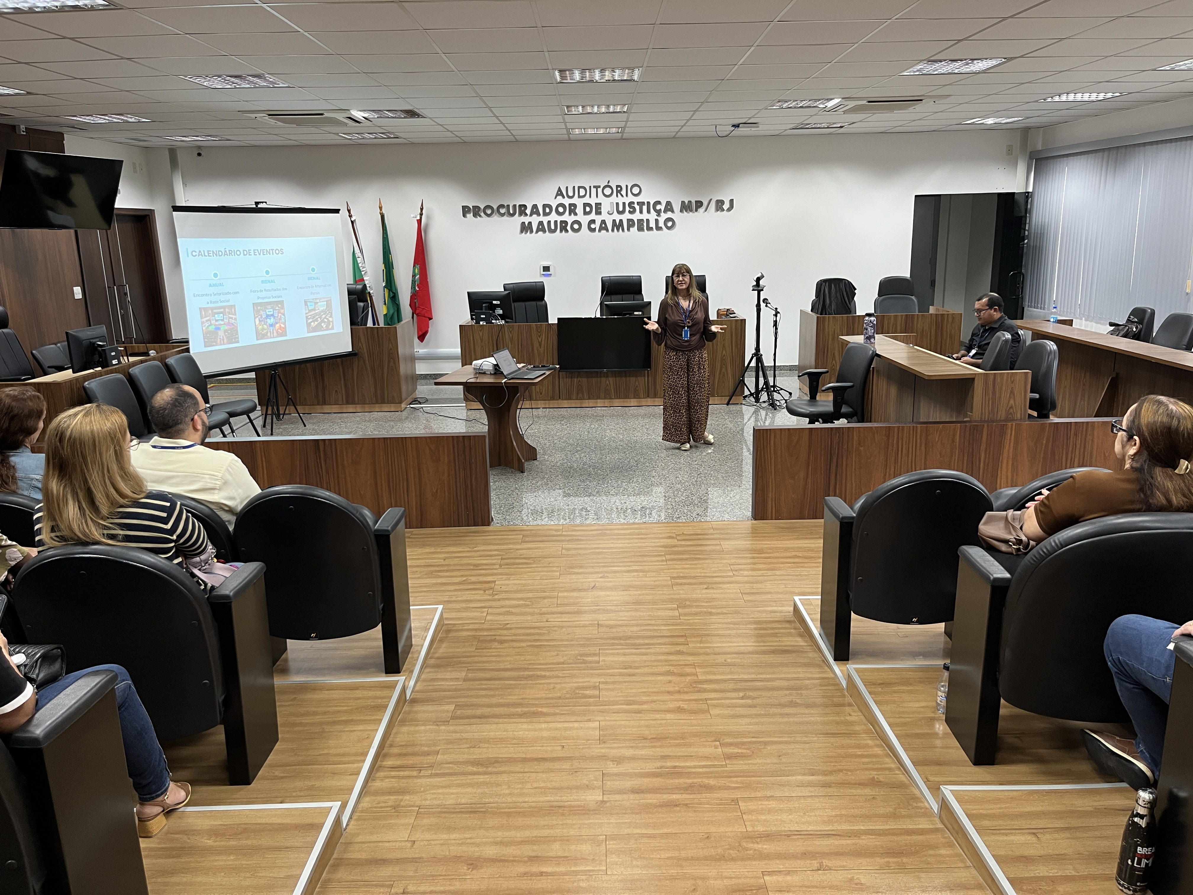 No mesmo auditório, Shirlene Fraxe está de pé, no centro do palco, com os braços levemente abertos, em um gesto de fala. Ela é uma mulher de cabelos castanhos, veste uma blusa marrom de mangas longas e uma saia longa com estampa animal print. À esquerda, uma tela de projeção exibe um slide com o título "Calendário de Eventos". O público acompanha a apresentação sentado nas poltronas.