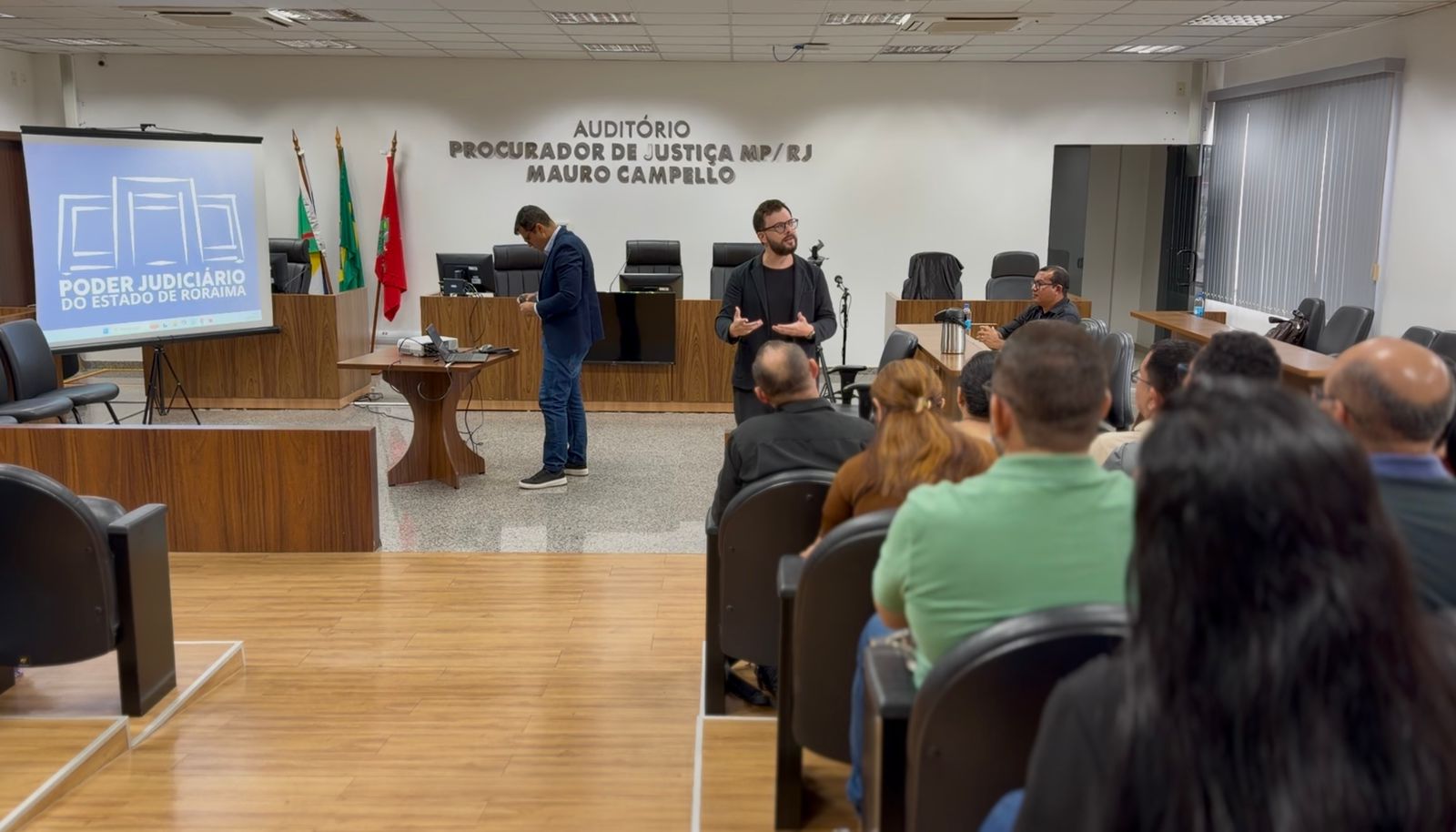 No mesmo auditório, o juiz Esdras Benchimol está de pé, gesticulando enquanto fala. Ele usa óculos, barba e veste um paletó cinza sobre uma camiseta preta. Ao seu lado, à esquerda, um homem de paletó azul e calça jeans organiza equipamentos sobre uma mesa de madeira. À esquerda, um projetor exibe o logotipo do Poder Judiciário do Estado de Roraima. O público, composto por diversas pessoas, assiste sentado em cadeiras estofadas escuras.