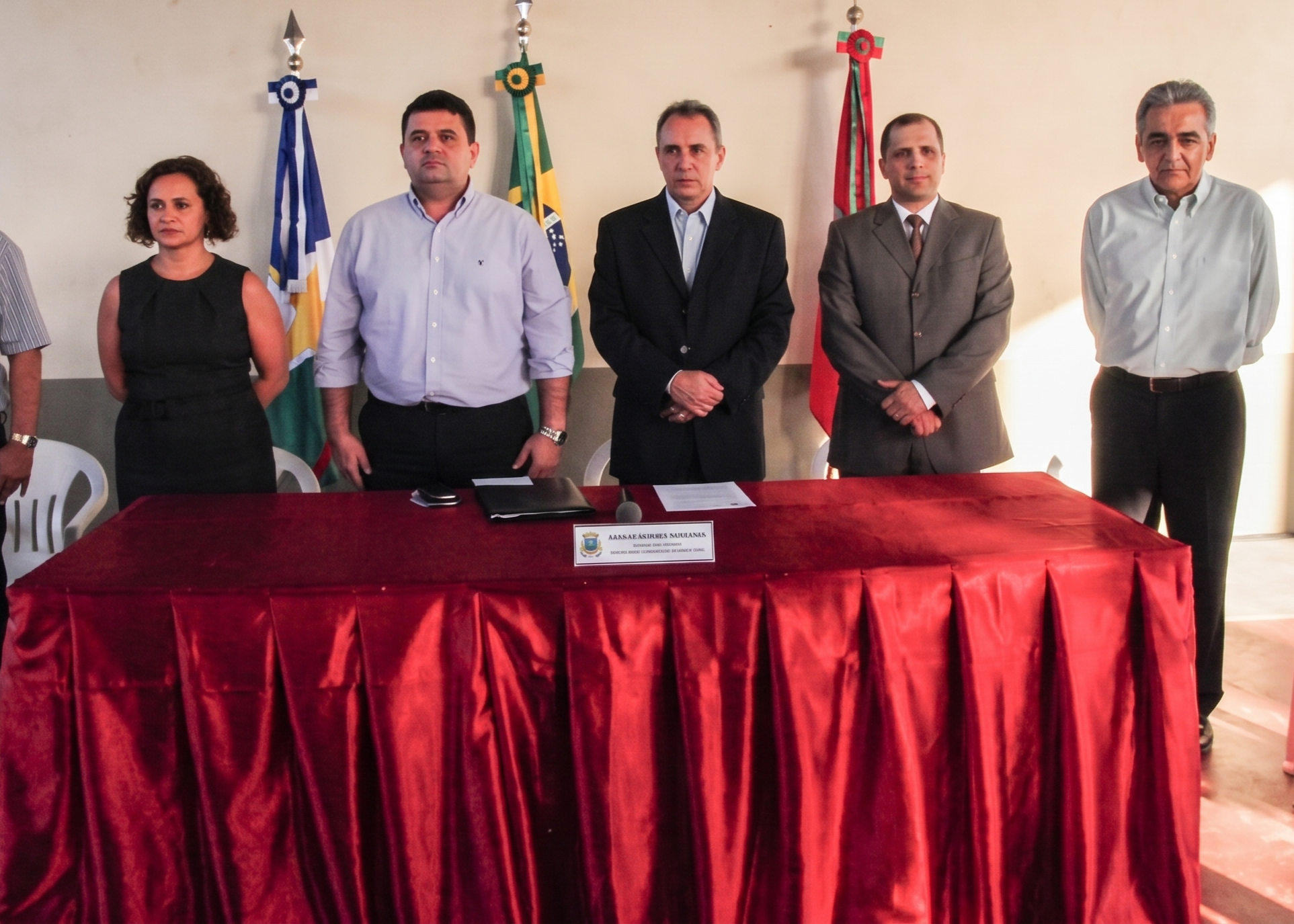 Imagem de arquivo colorida mostra autoridades em pé atrás de uma mesa coberta com tecido vermelho, durante cerimônia institucional. Ao fundo, aparecem bandeiras e uma parede clara.