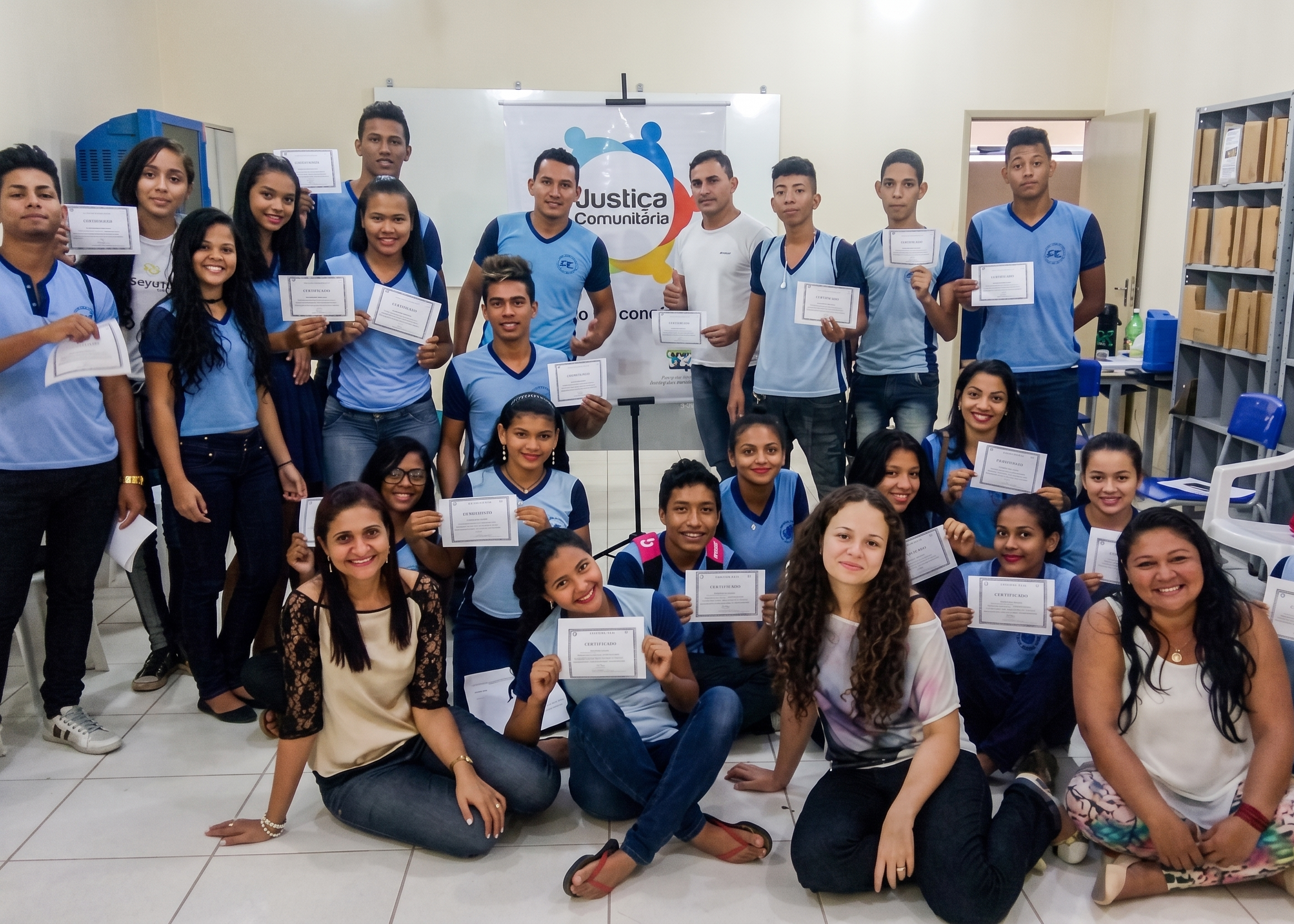 Foto colorida mostra estudantes e participantes posando em grupo e segurando certificados. Ao fundo, há um banner com a identidade visual da Justiça Comunitária.