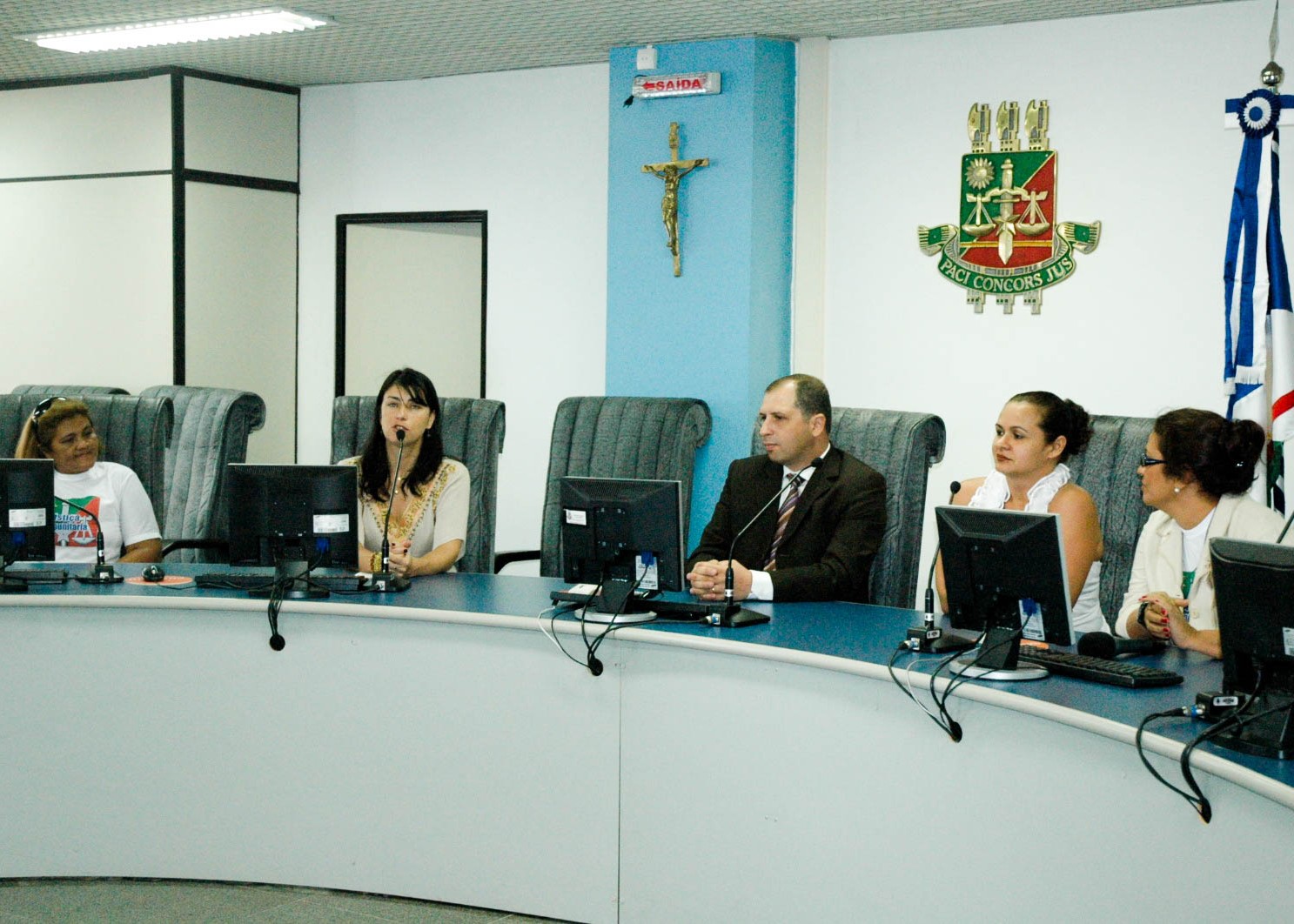 Foto colorida mostra reunião em uma sala do Tribunal de Justiça de Roraima. Participantes estão sentados em uma bancada com microfones e computadores, durante atividade ligada à Justiça Comunitária.