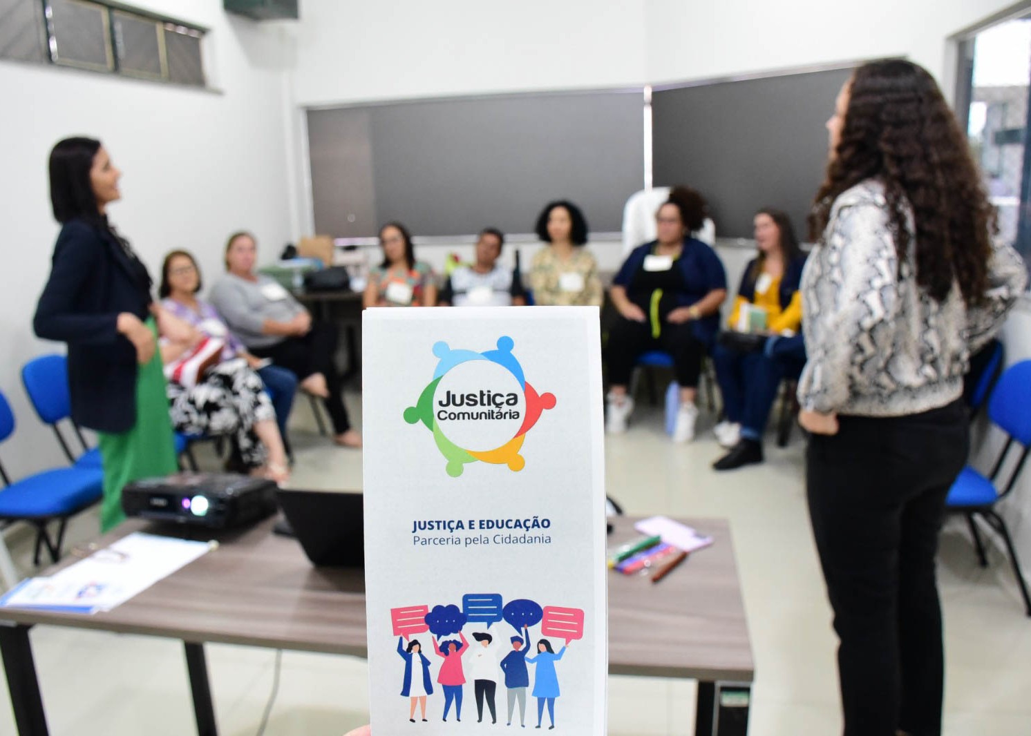 Foto colorida mostra, em primeiro plano, um material informativo da Justiça Comunitária com o tema “Justiça e Educação: Parceria pela Cidadania”. Ao fundo, pessoas participam de uma roda de conversa em uma sala.