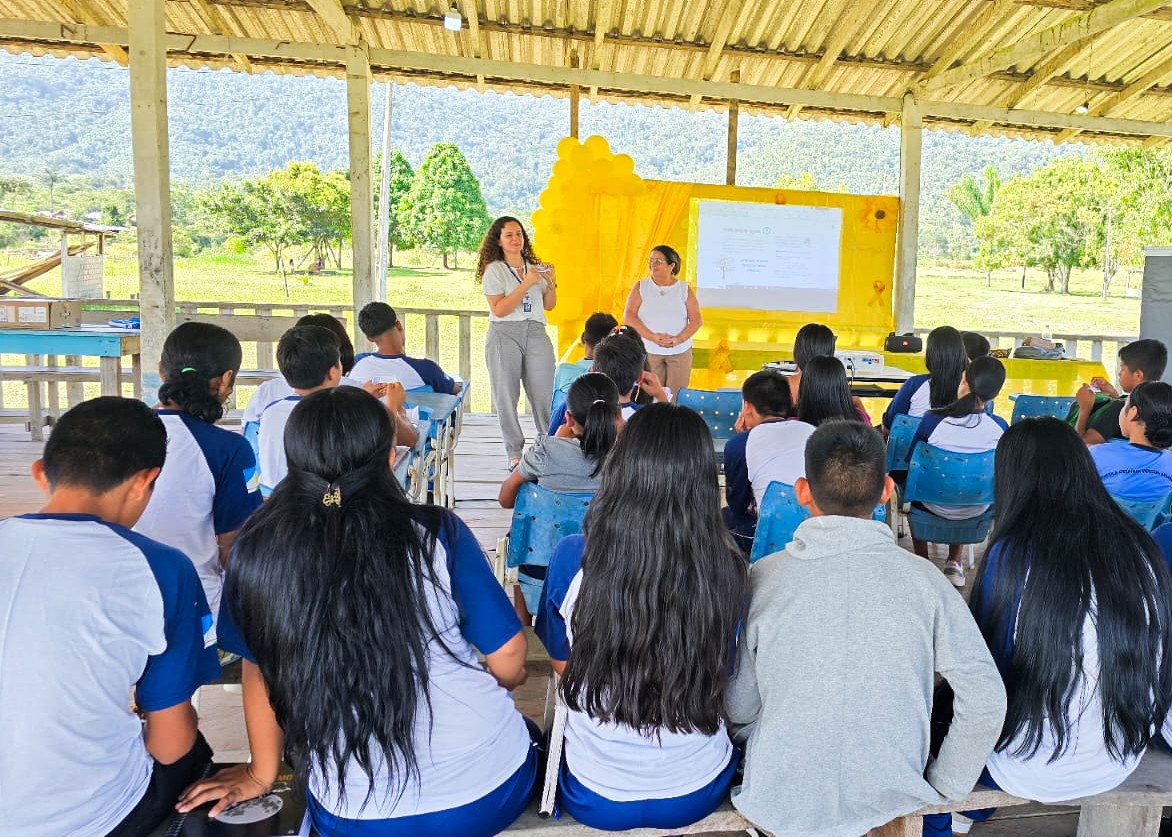 Foto colorida mostra estudantes sentados em uma escola durante palestra sobre violência. À frente, duas mulheres conduzem a atividade, com apoio de uma apresentação projetada em uma tela.