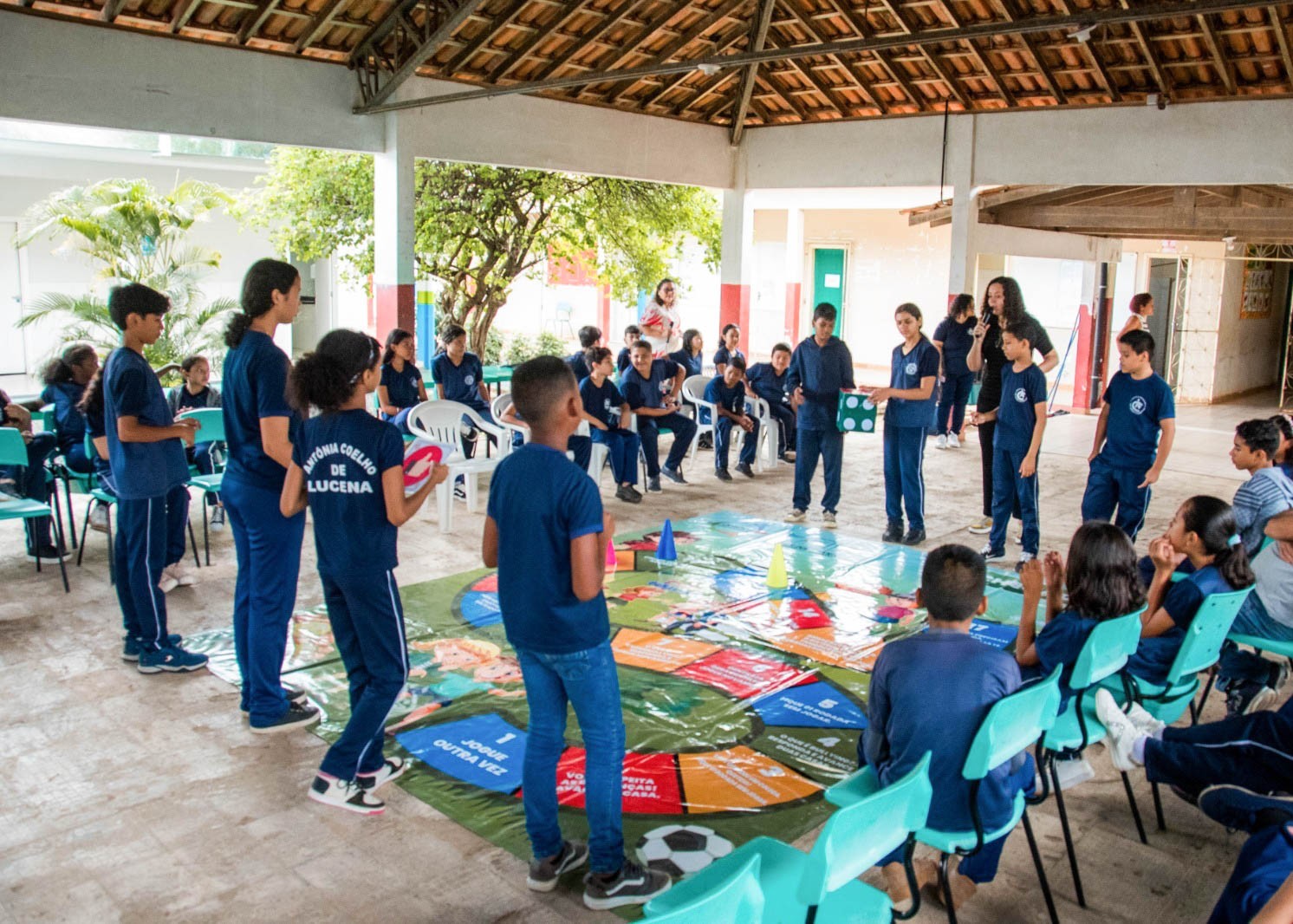 Foto colorida mostra estudantes participando de uma atividade com tabuleiro gigante no chão. Crianças e adolescentes estão em roda, acompanhando a dinâmica conduzida por uma mulher ao microfone.