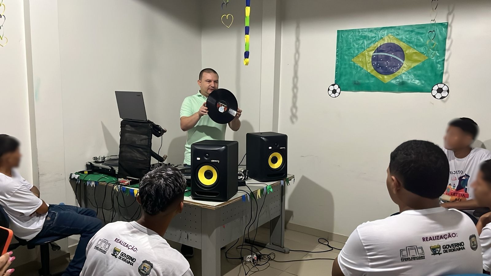 Homem apresentando um disco de vinil em uma oficina, com equipamento de som ao fundo e bandeira do Brasil na parede. Pessoas participam da atividade. Homem apresentando um disco de vinil em uma oficina, com equipamento de som ao fundo e bandeira do Brasil na parede. Pessoas participam da atividade.