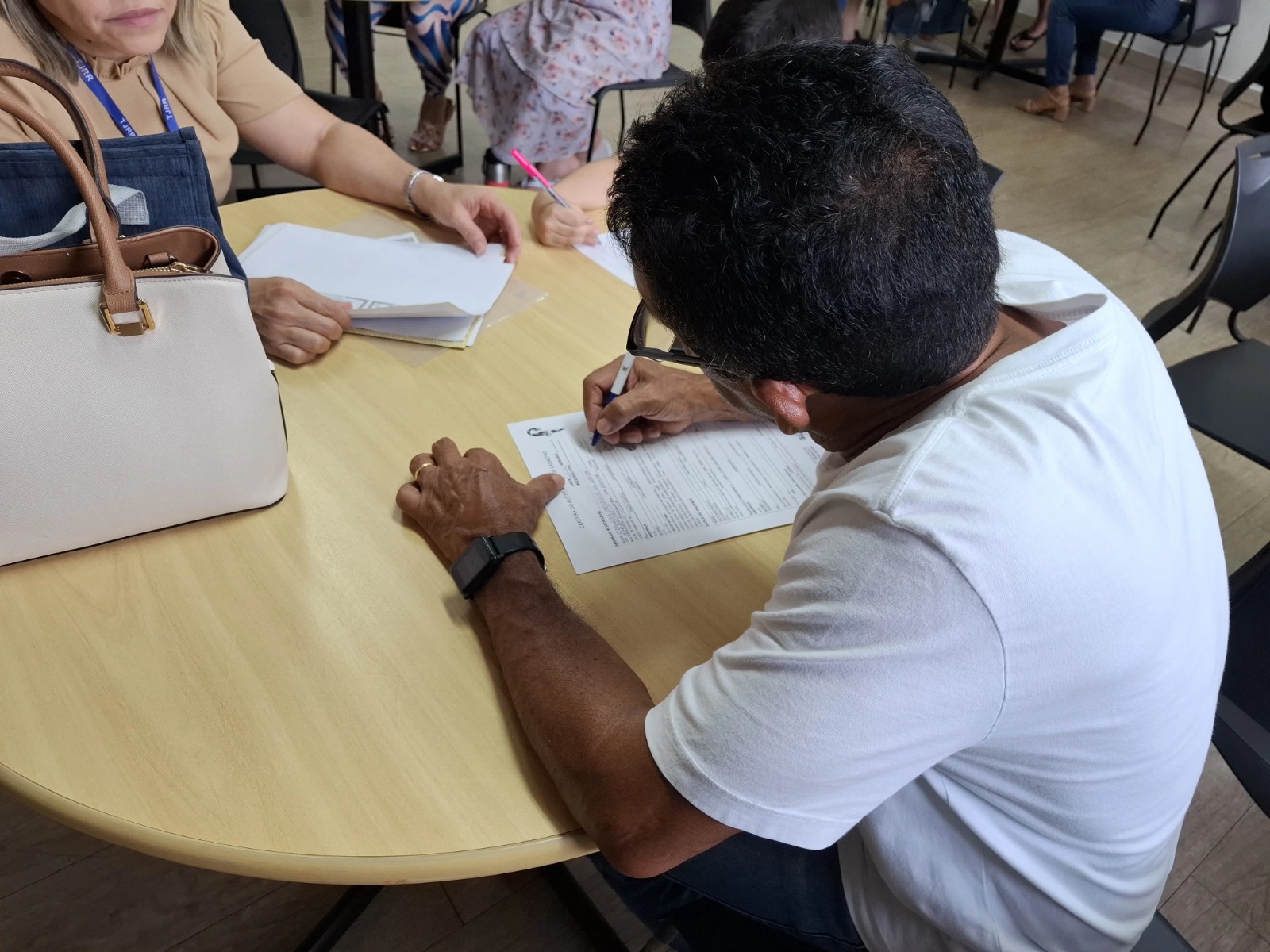 Homem escrevendo em um formulário em uma mesa, com uma mulher ao fundo segurando papéis. Uma bolsa está sobre a mesa. Homem escrevendo em um formulário em uma mesa, com uma mulher ao fundo segurando papéis. Uma bolsa está sobre a mesa.