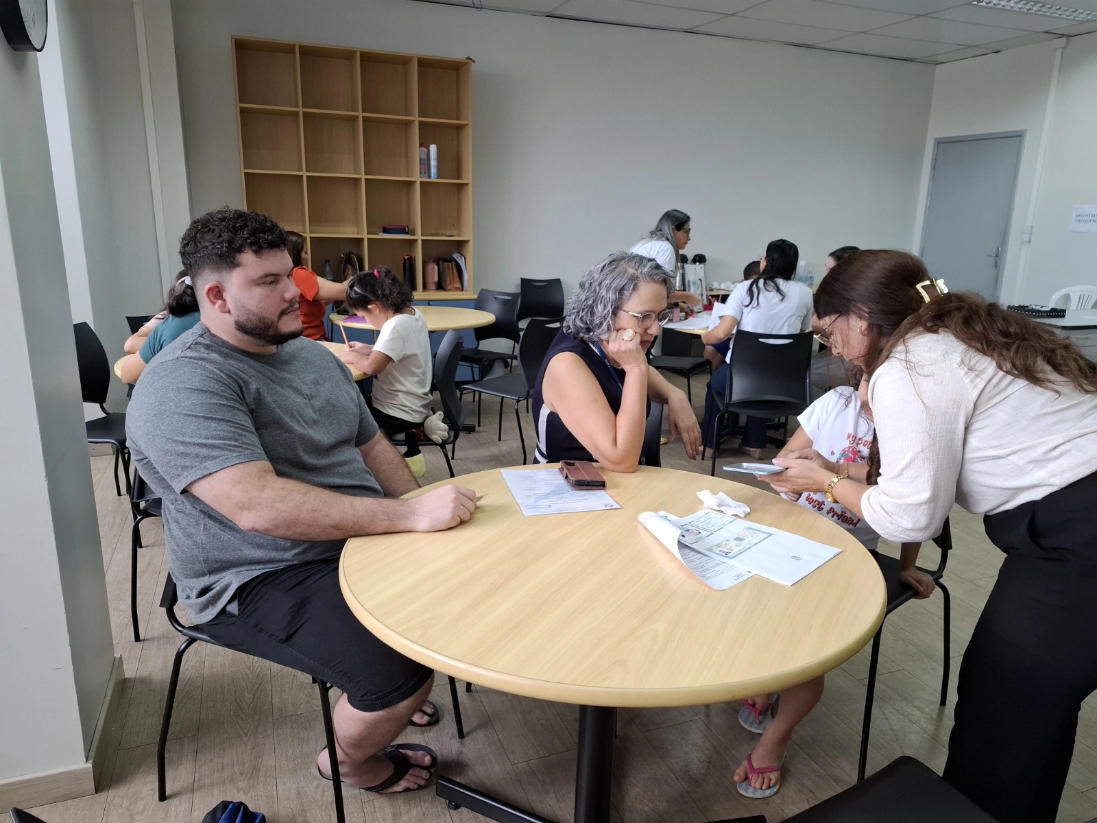 Grupo de pessoas em uma sala, algumas sentadas em uma mesa redonda, conversando e usando dispositivos móveis. Grupo de pessoas em uma sala, algumas sentadas em uma mesa redonda, conversando e usando dispositivos móveis.