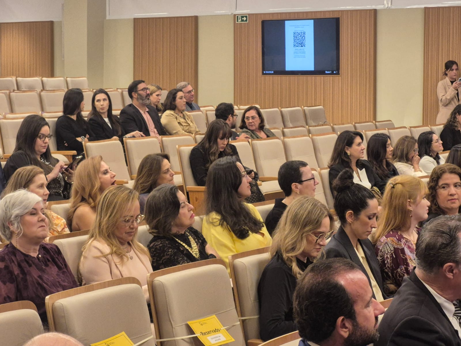 Imagem colorida mostra uma visão em ângulo de um auditório com várias pessoas sentadas em poltronas beges, destacando-se entre elas a juíza Suelen Alves, vestindo amarelo, focada no evento. Imagem colorida mostra uma visão em ângulo de um auditório com várias pessoas sentadas em poltronas beges, destacando-se entre elas a juíza Suelen Alves, vestindo amarelo, focada no evento.